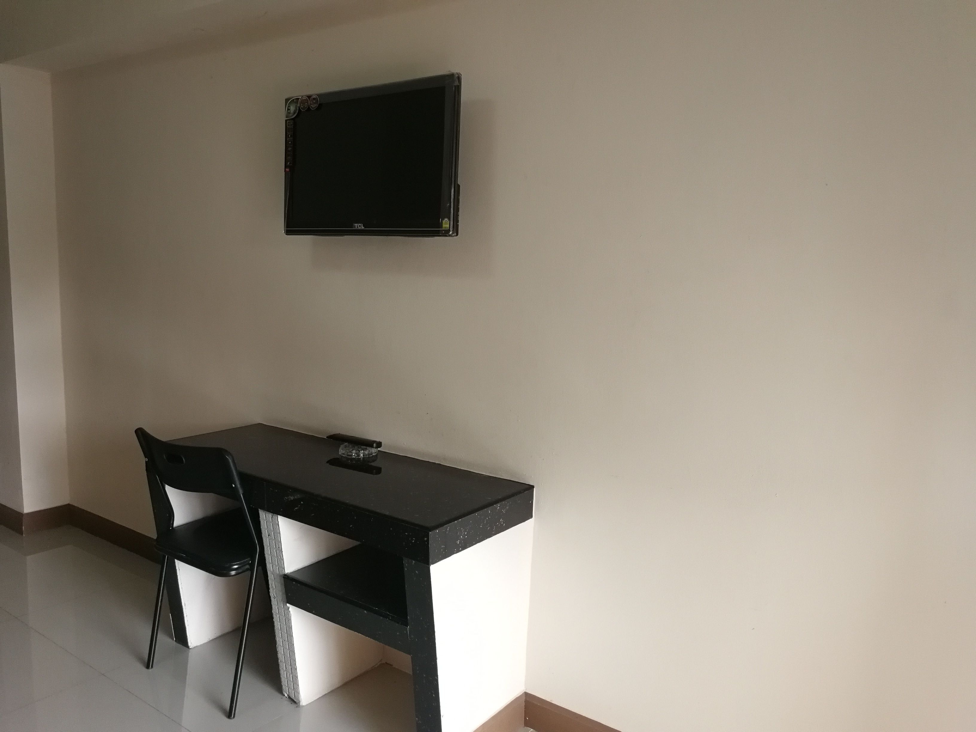 Una habitación pequeña y sencilla con escritorio y silla, un televisor de pantalla plana montado en la pared, y un estante pequeño.