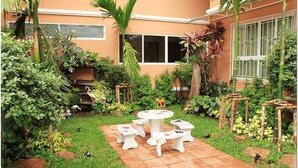 Eine Terrasse mit rundem Tisch und Bänken, umgeben von üppigem Grün und einem gefliesten Boden.