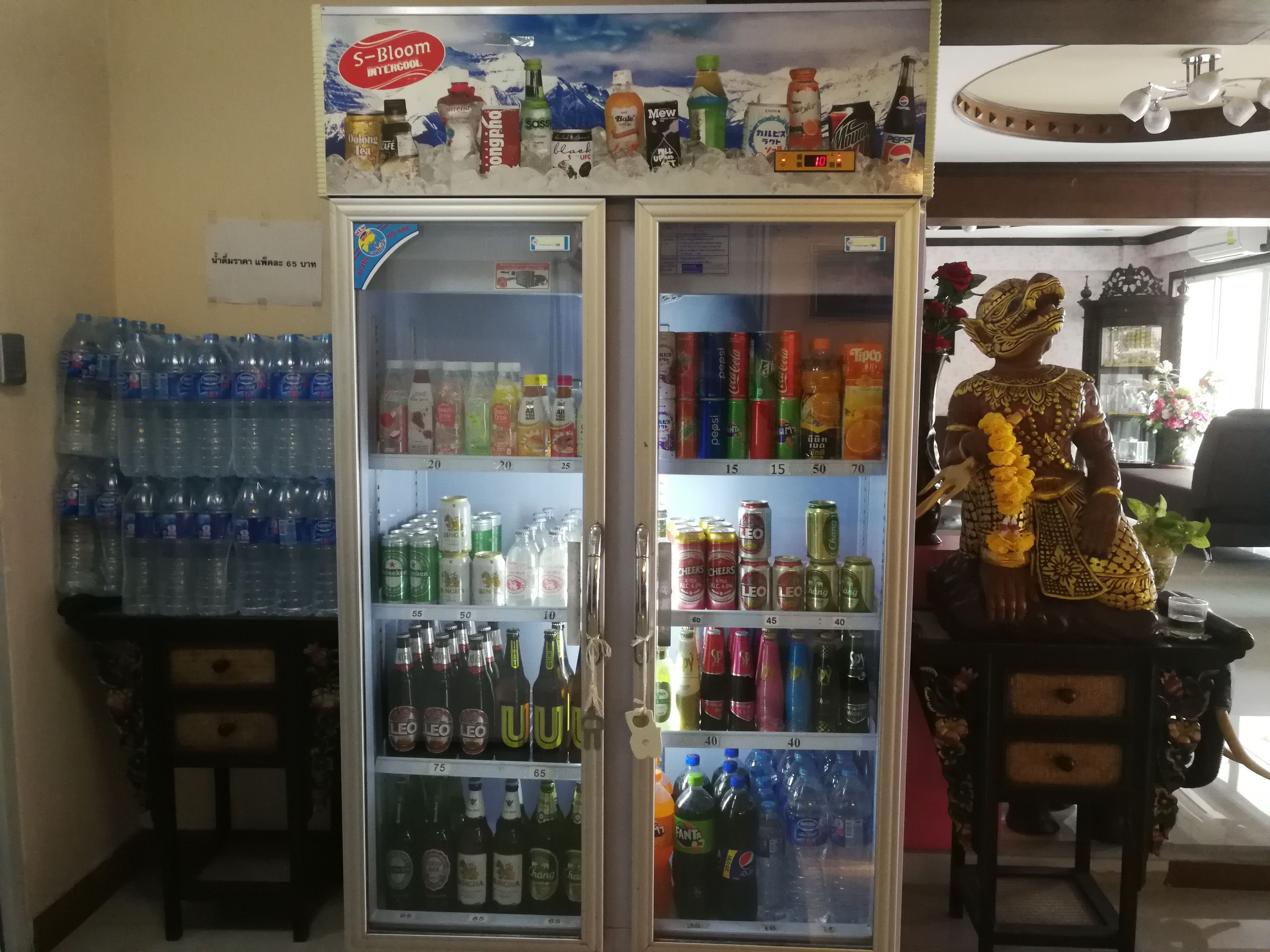 Un refrigerador para bebidas repleto de diversas opciones, una figura decorativa y botellas de agua en estantes.