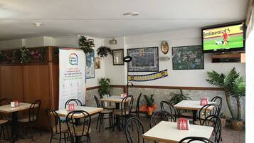 Un restaurante con varias mesas y sillas, un cartel de menú, un televisor montado en la pared y varias decoraciones, incluyendo cuadros enmarcados y un banderín.