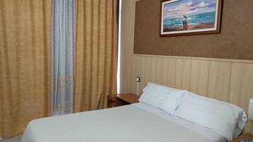 Una habitación de hotel con cama, mesita de noche, un cuadro de una playa y una ventana con cortinas.