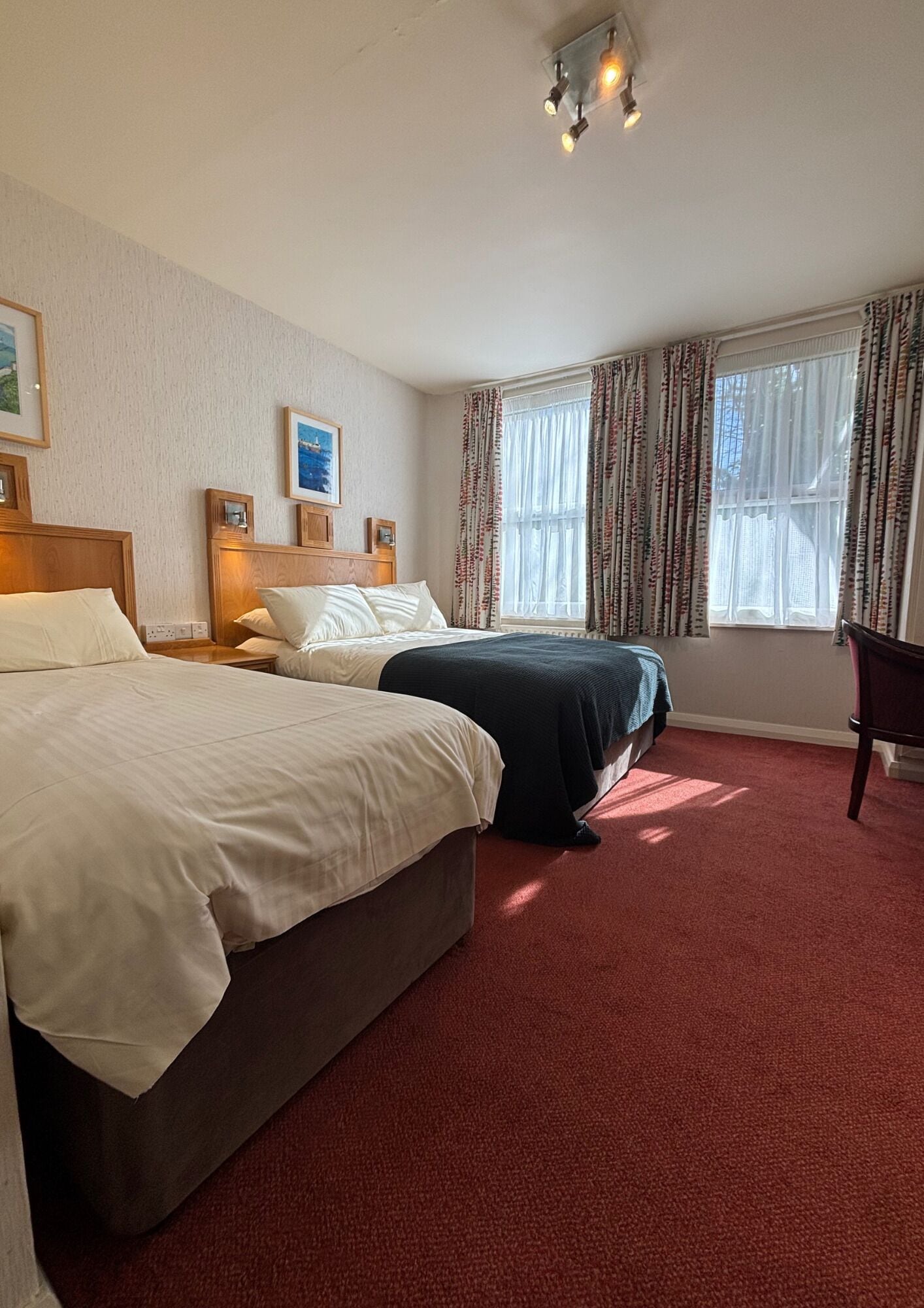 A hotel room with two beds, a red carpet, a wooden headboard, a desk, and a chair.