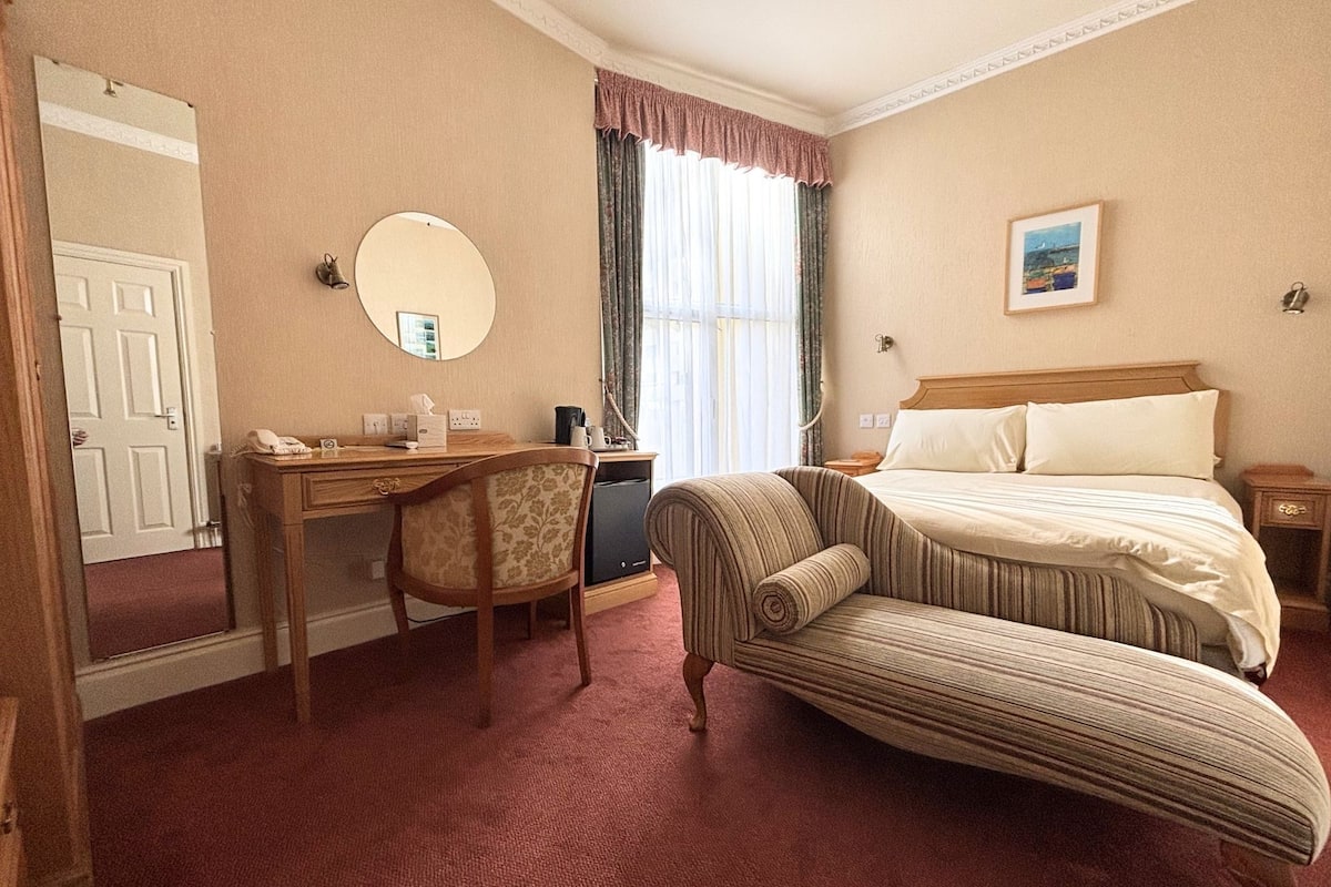 Une chambre d’hôtel avec un lit, un bureau, une chaise, un miroir et une fenêtre avec des rideaux.