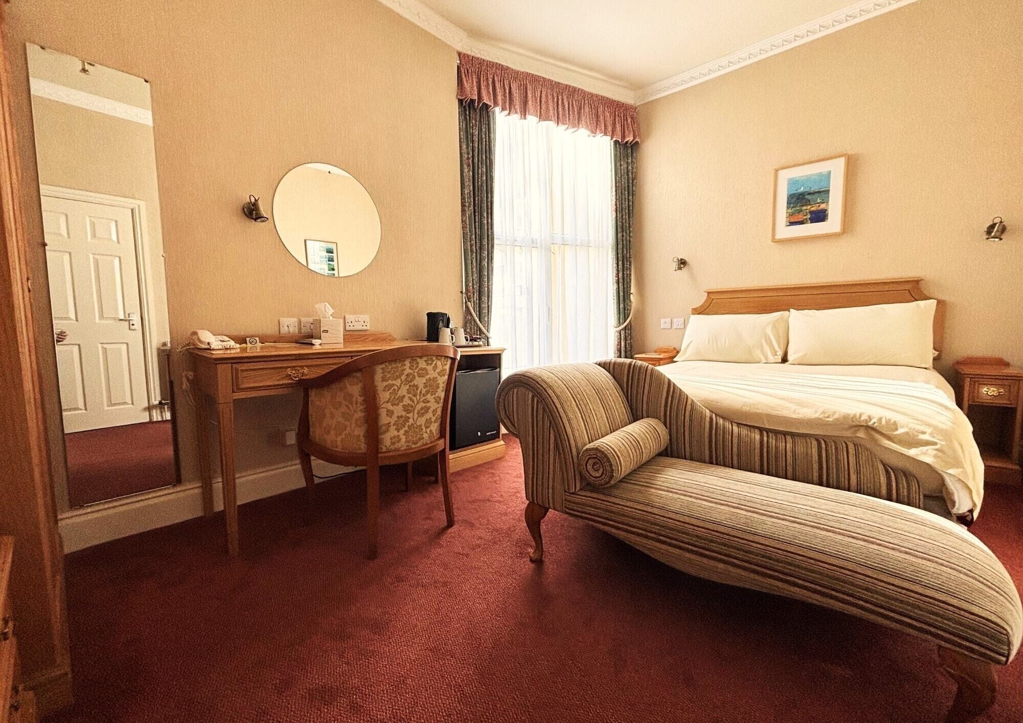 A hotel room with a bed, a desk, a chair, a mirror, a window with curtains, and a painting on the wall.