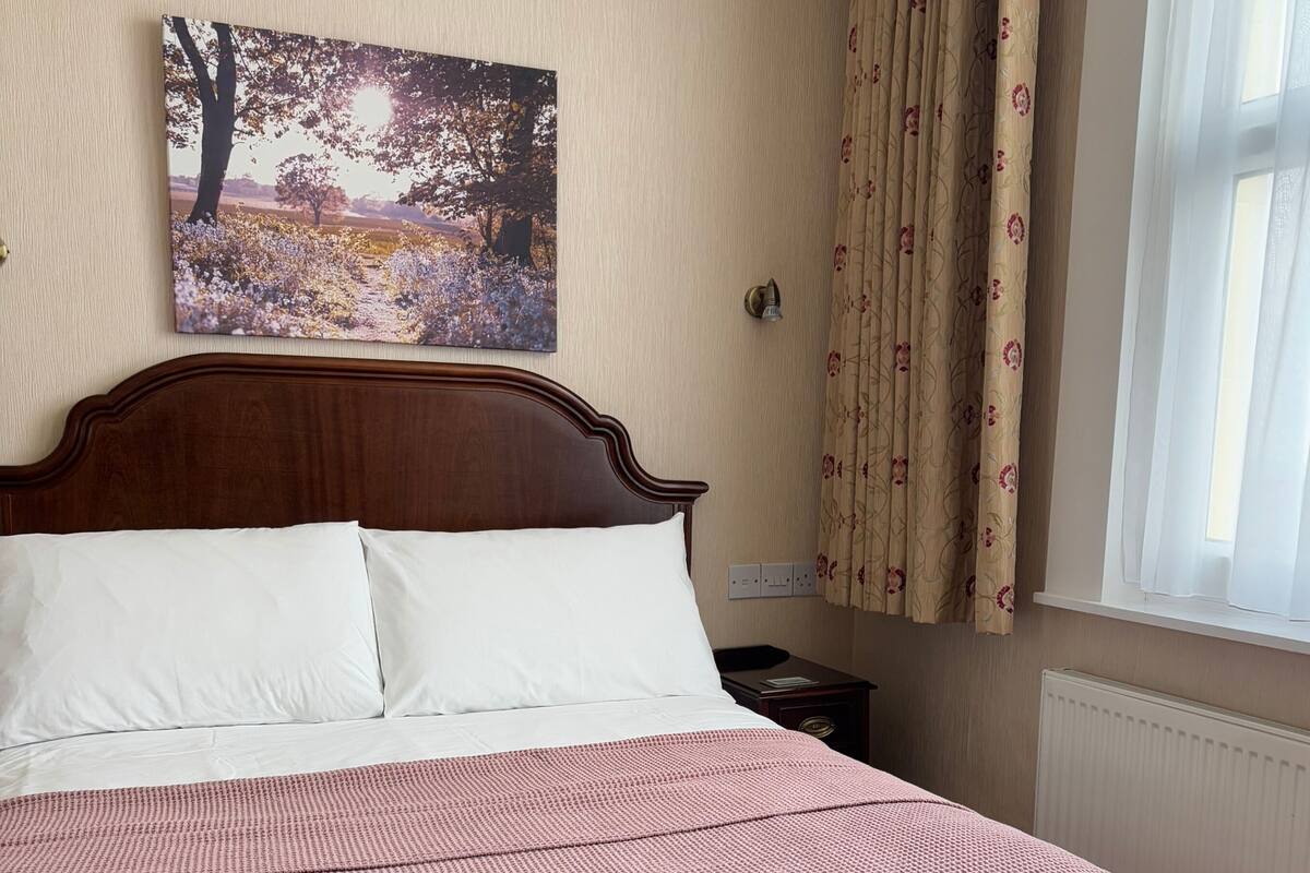 Un lit bien fait, recouvert d’une couvre-lit à carreaux, avec une tête de lit en bois, un tableau accroché au mur, une table de chevet avec une lampe et une fenêtre avec des rideaux.