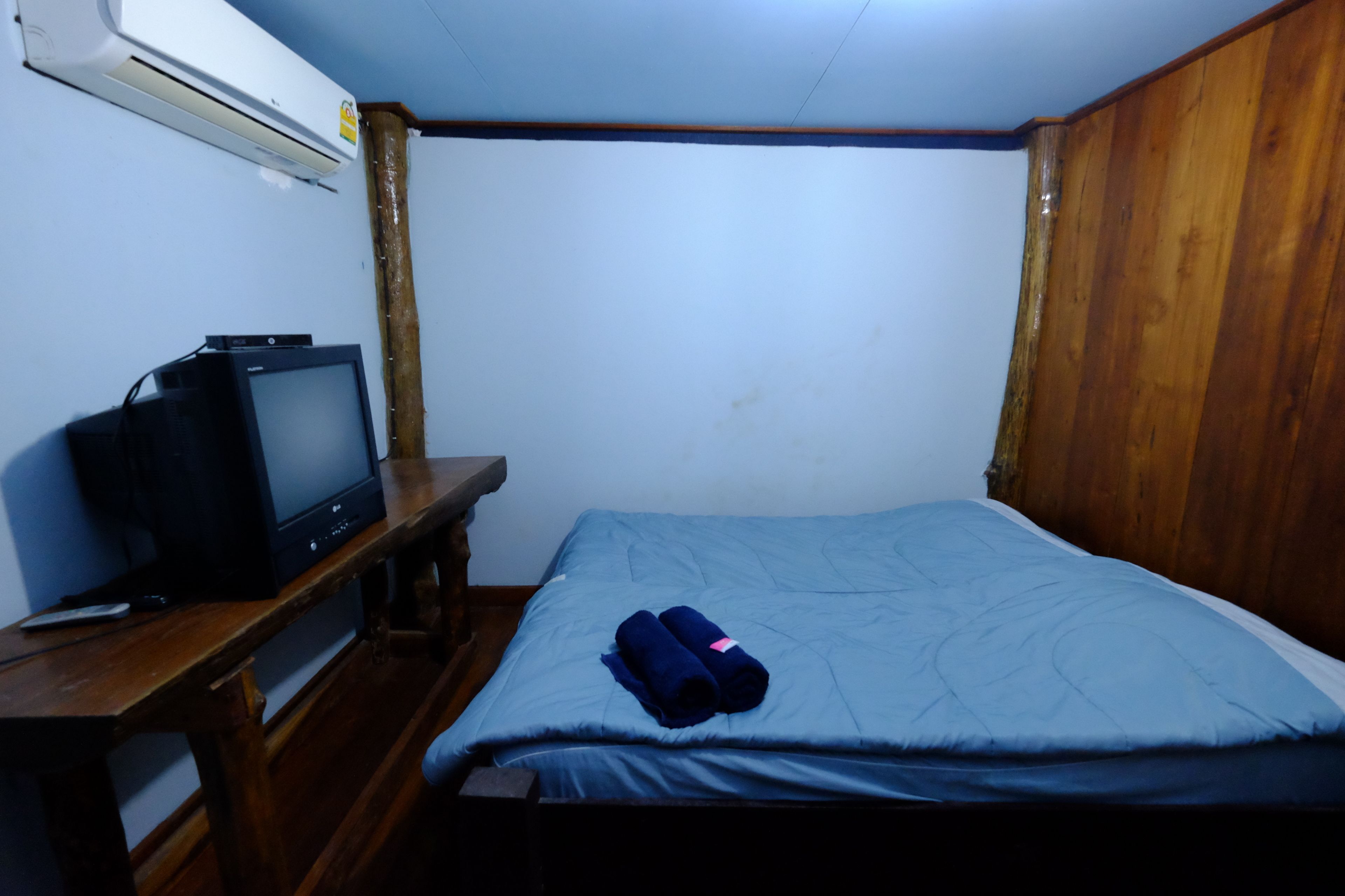 A small room with a bed, a wooden desk, a television, and a wall-mounted air conditioner.