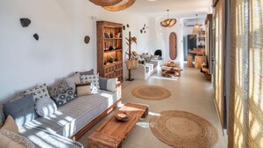A modern living room with a sofa, coffee table, and decorative items. There is a bookshelf and a dining area in the background. - Ayama Hideaway Villa by Calm Collection (Santorini)