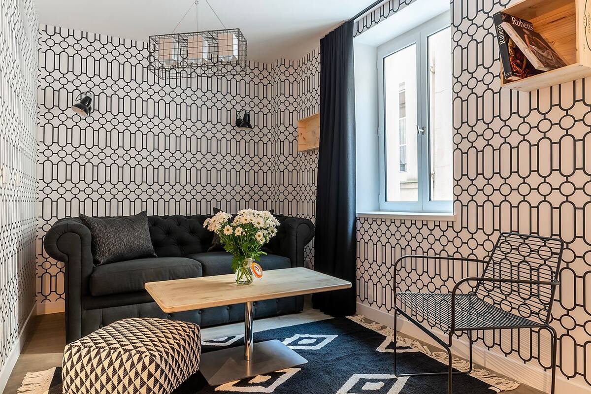 Ein modernes Wohnzimmer mit geometrischer Wandtapete, einer schwarzen Couch, einem Holztisch mit Blumenvase, einem gemusterten Hocker und einem Metallstuhl.