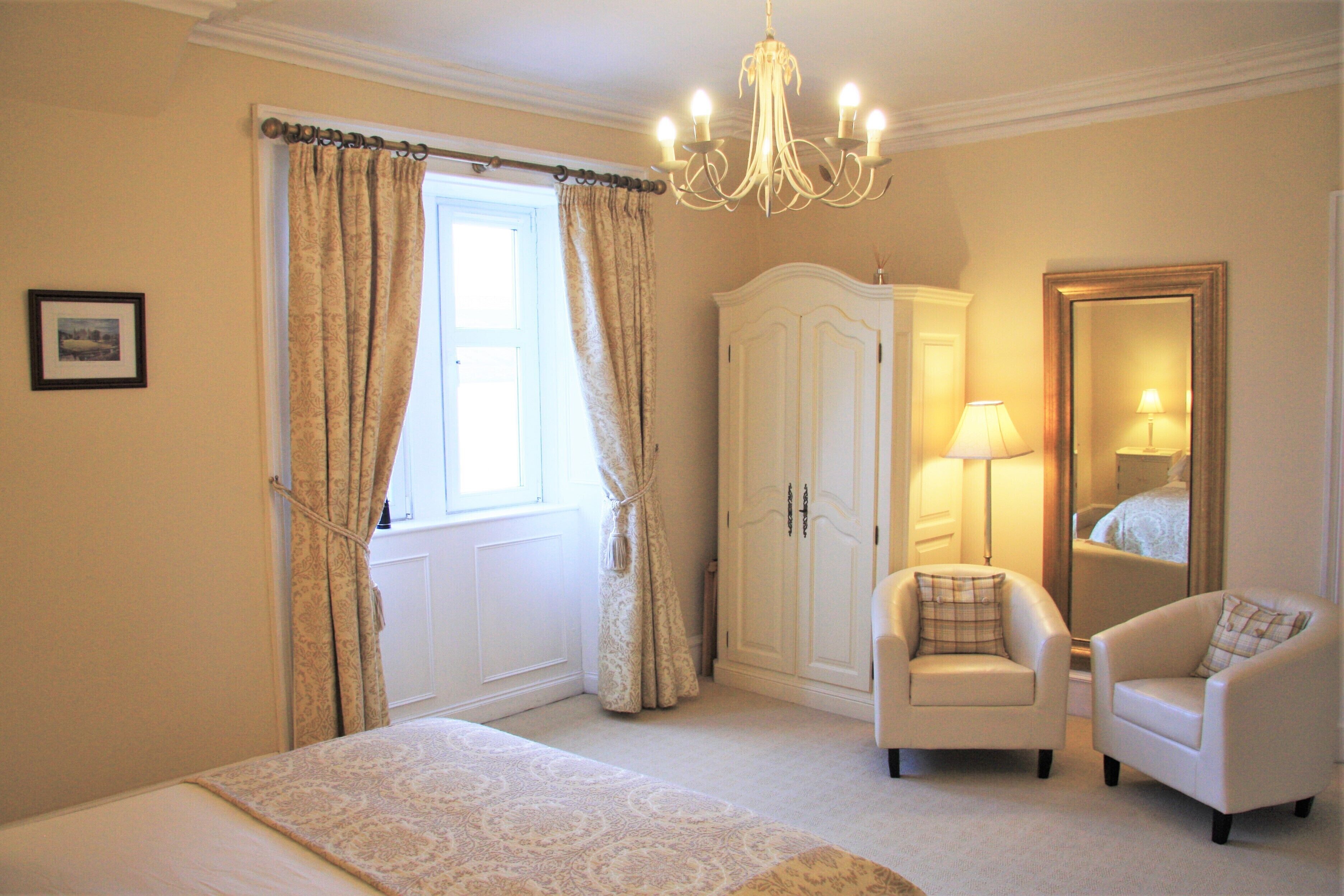 A bedroom with a bed, two chairs, a wardrobe, a chandelier, and a mirror.