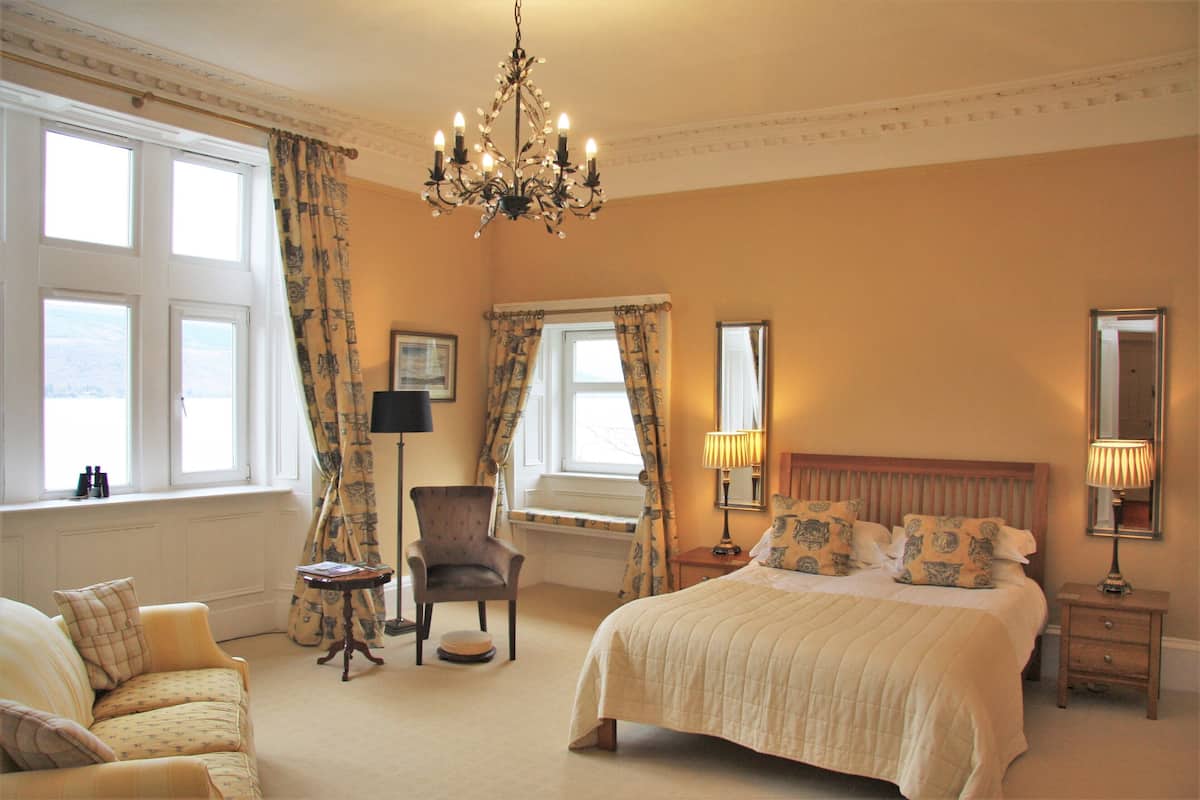 A hotel room with a large bed, a sitting area with a chair and table, a chandelier, and large windows with curtains.