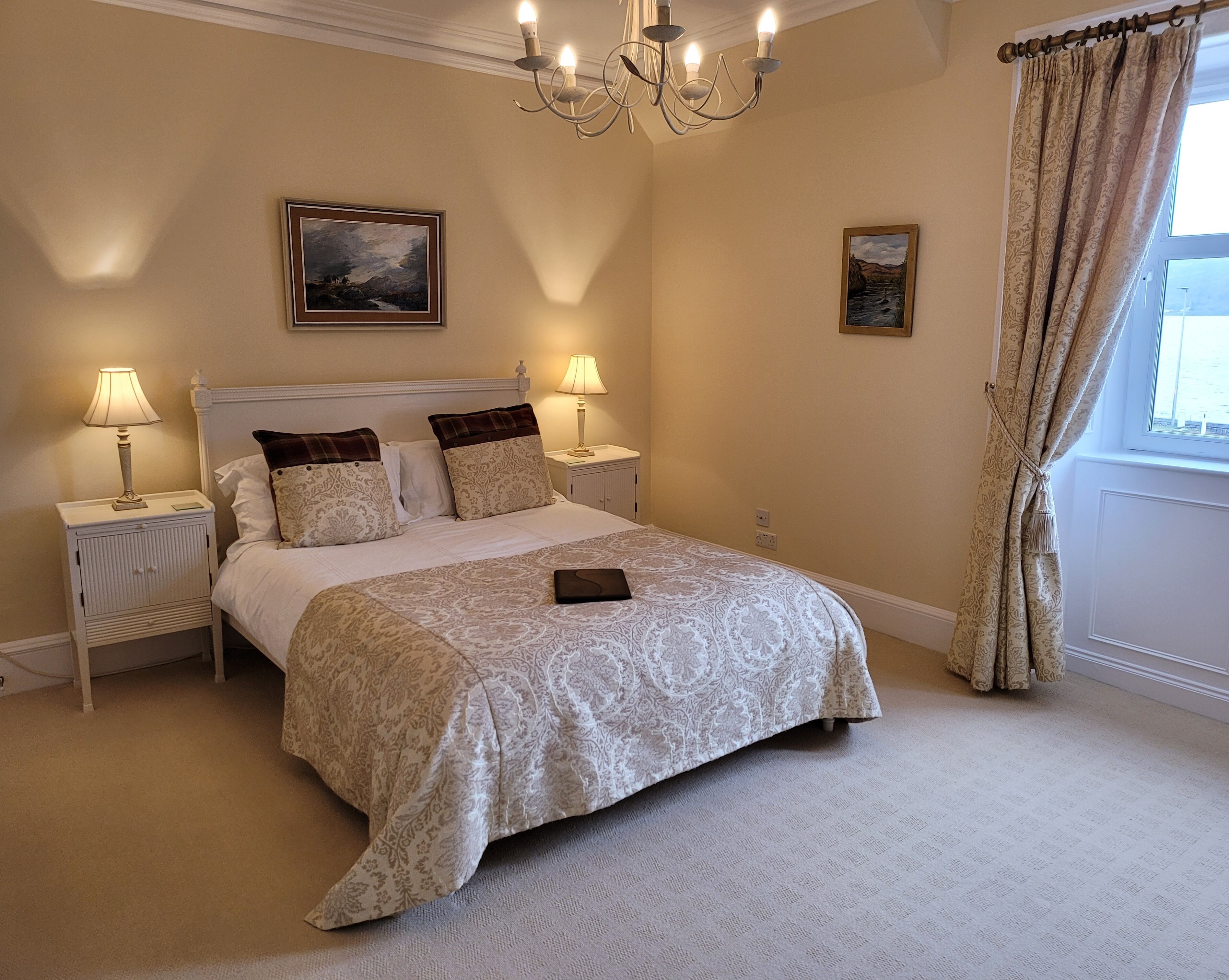 A neatly arranged bedroom with a large bed, two bedside tables with lamps, a chandelier, and two framed pictures on the walls.