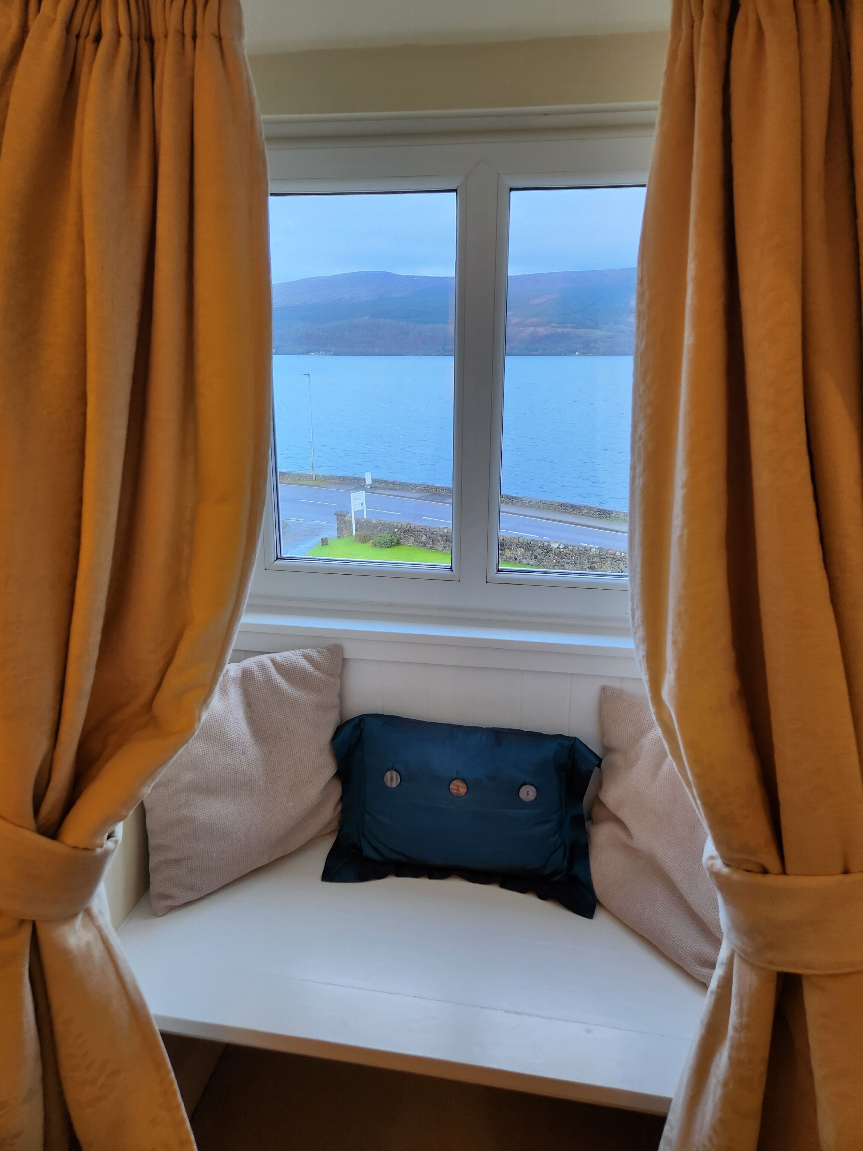 A window seat with a view of a lake and mountains, curtains, and a few pillows.