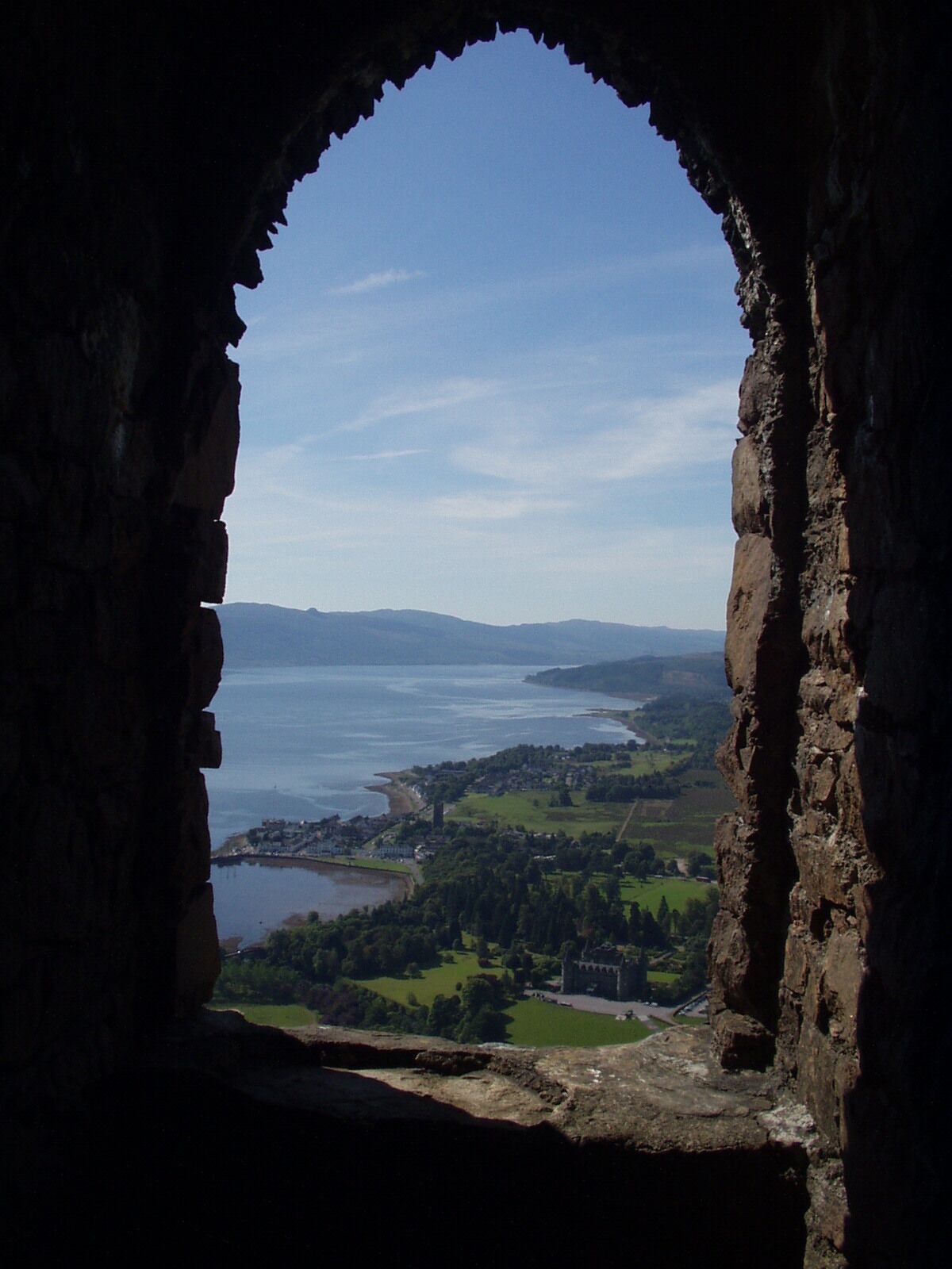 A view through an arched window of a coastal landscape with a body of water, green fields, and a distant town.
