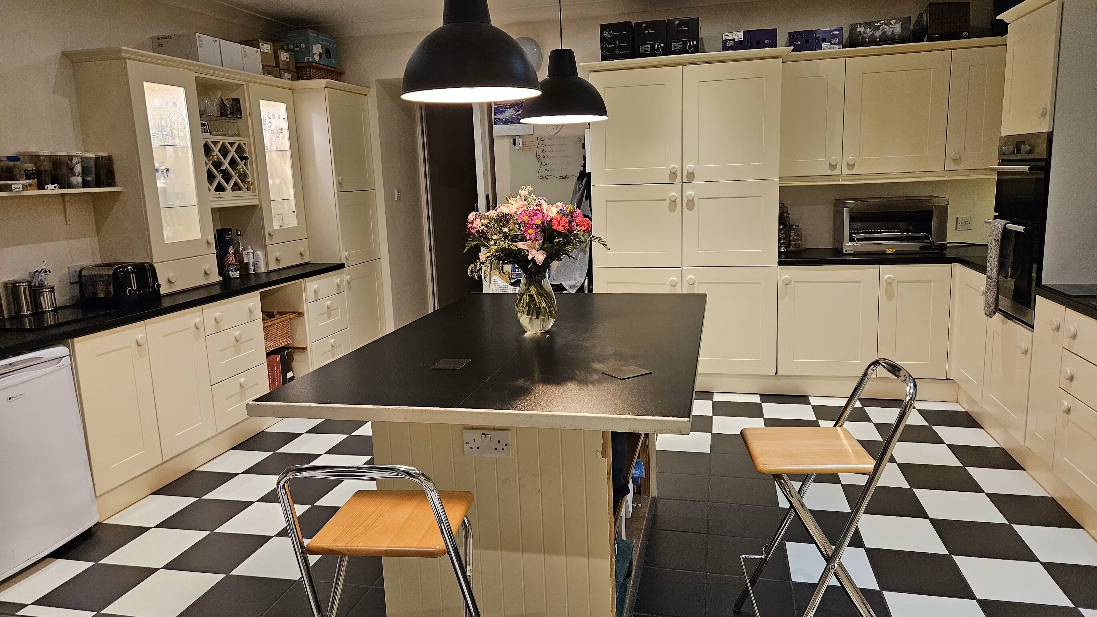 Uma cozinha com ilha central, duas cadeiras, um vaso com flores e piso xadrez.