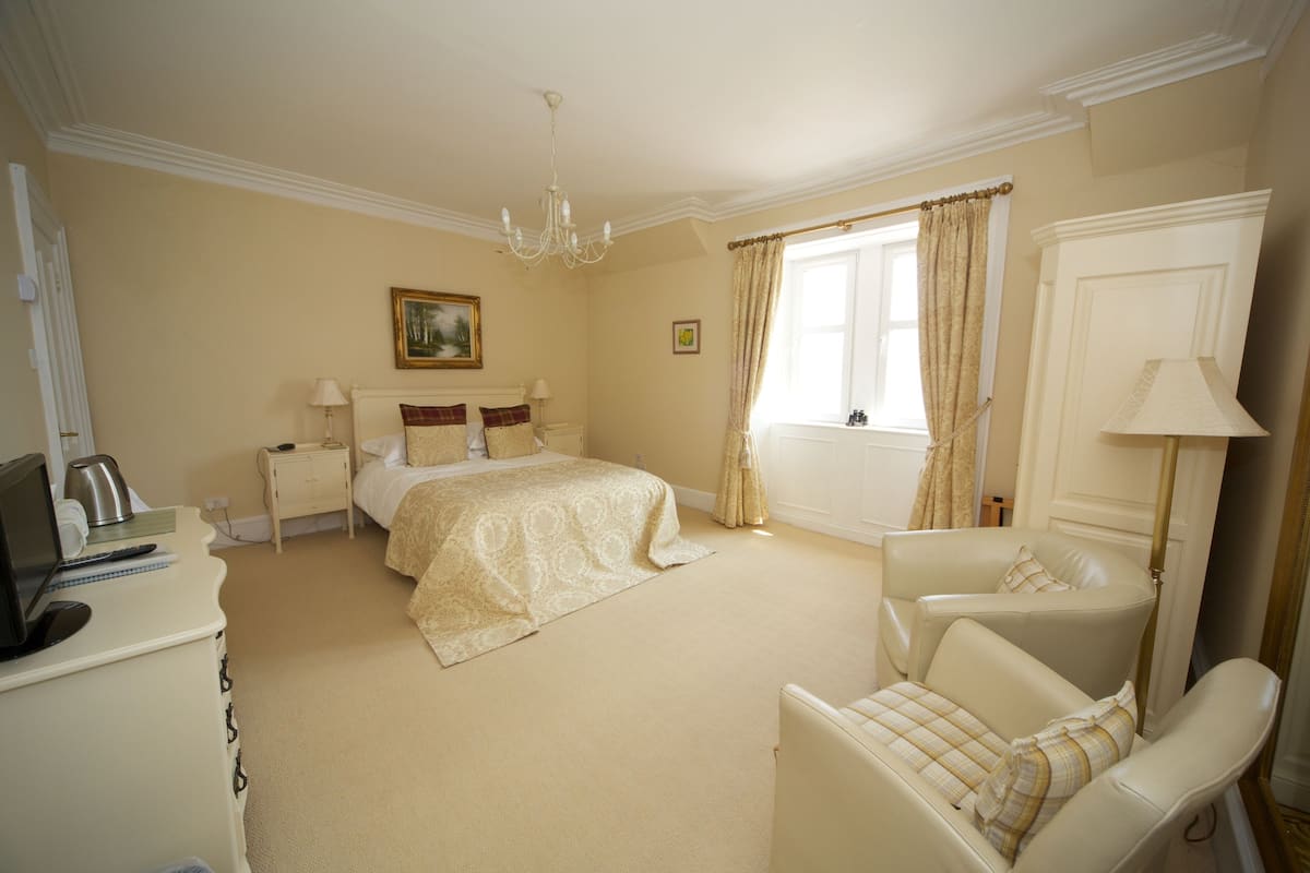 A hotel room with a bed, a desk with a computer, a chair, a window with curtains, and a chandelier.