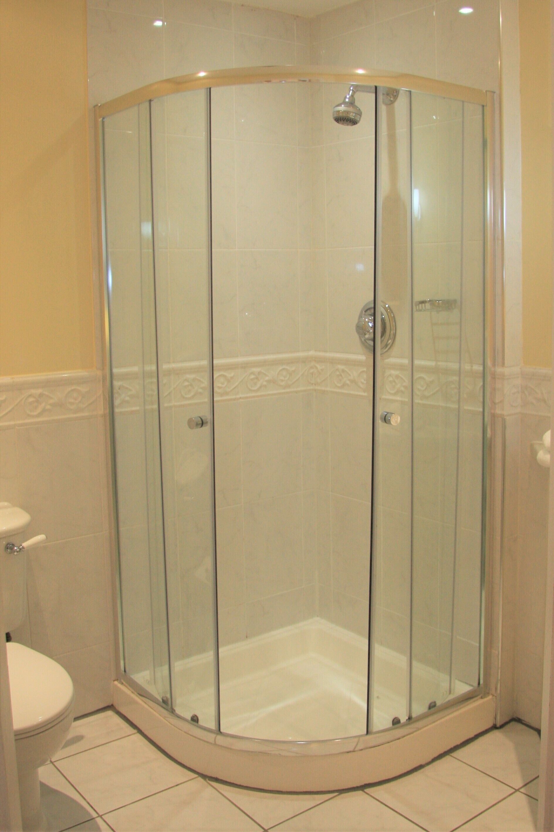 A glass-enclosed shower area with a white tiled floor and walls, a toilet to the left, and a showerhead above the tub.