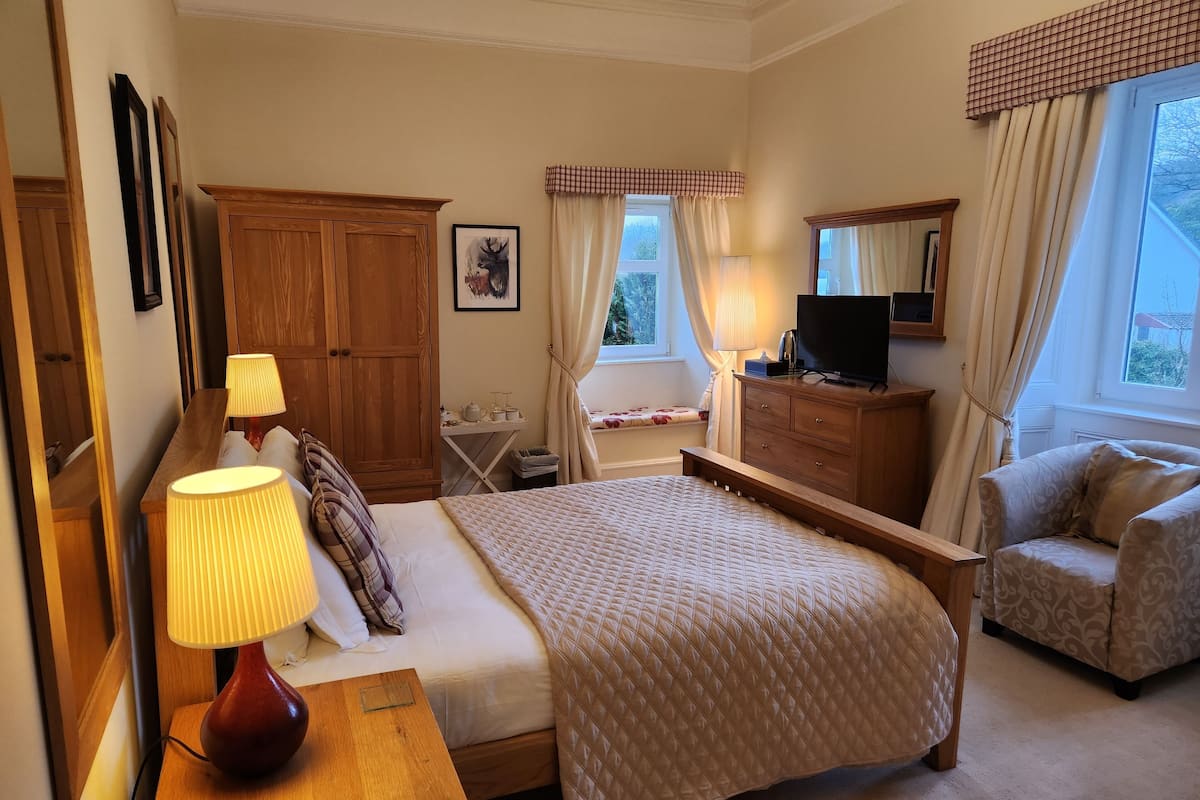 A neatly arranged bedroom with a bed, bedside table, lamp, wooden wardrobe, dresser, and a chair.
