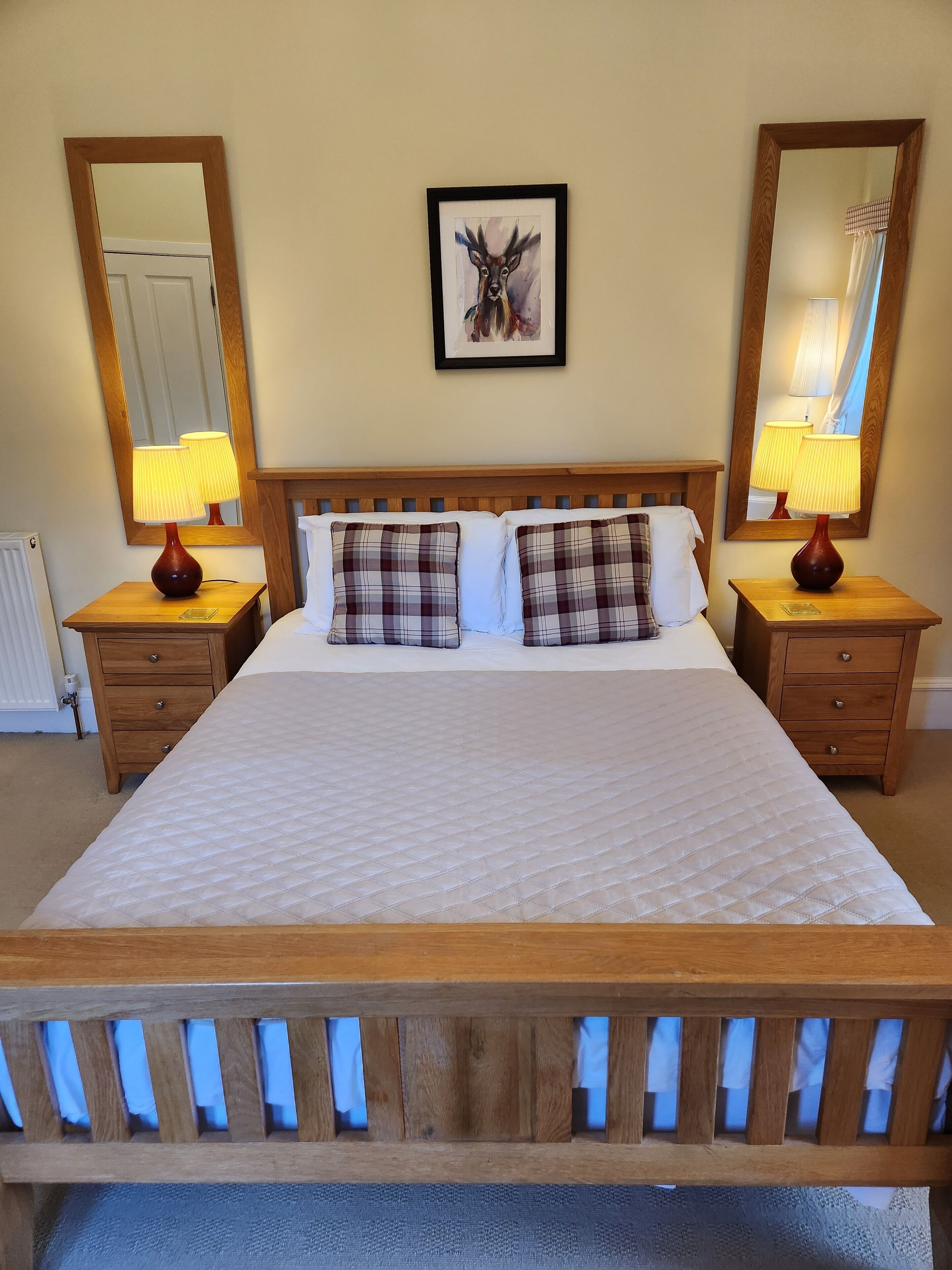A neatly made bed with a quilted white duvet, flanked by two bedside tables with lamps, a headboard with a mirror, and a framed artwork above the bed.