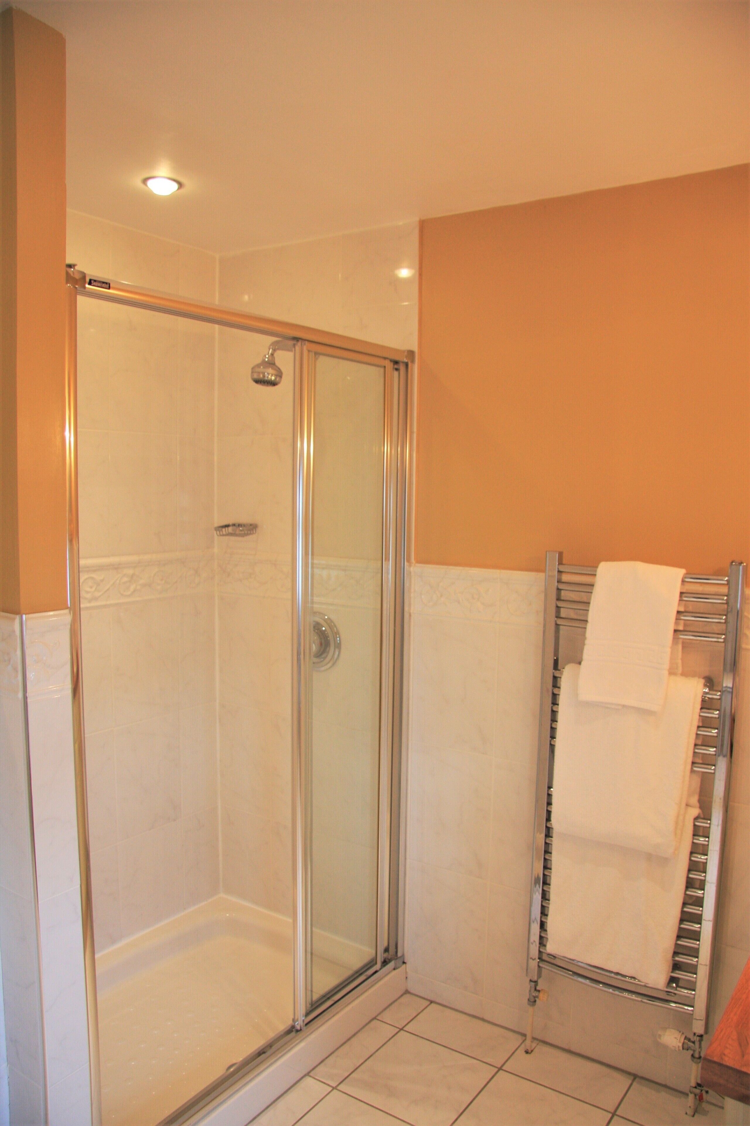 Ducha con paredes de vidrio, marco de cromo, pared de azulejos blancos y un perchero con una toalla blanca.