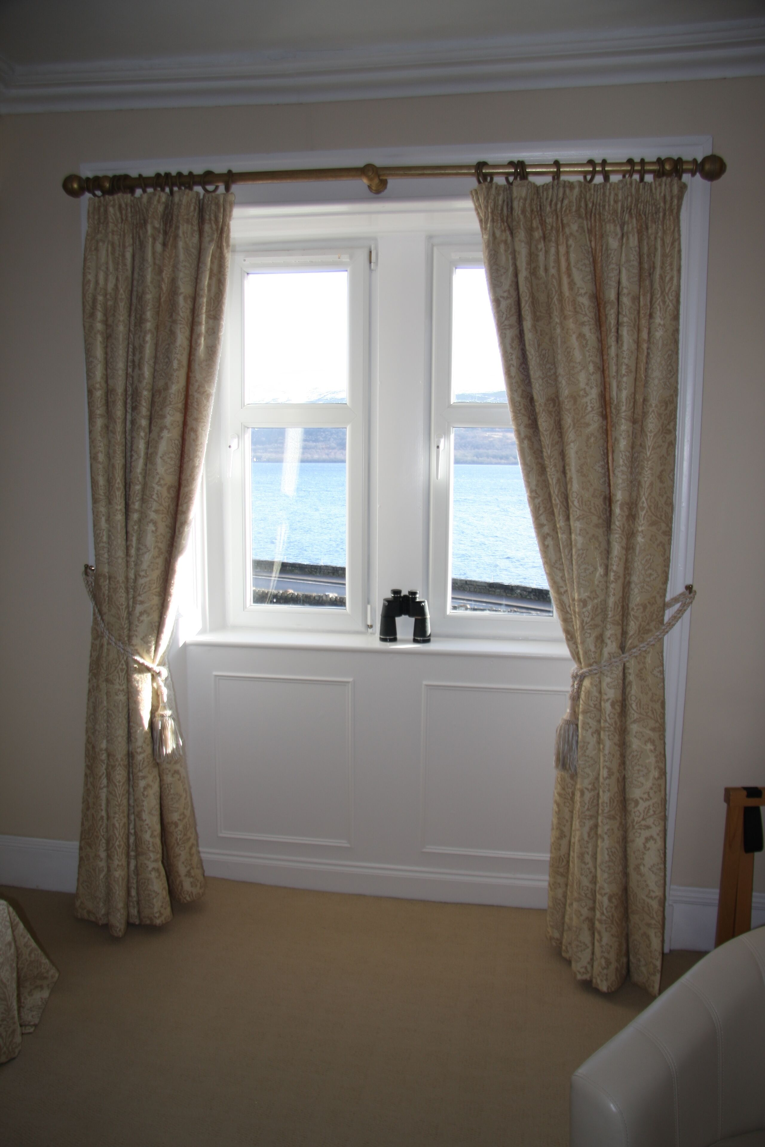 A room with a large window, beige curtains, and a view of the sea.