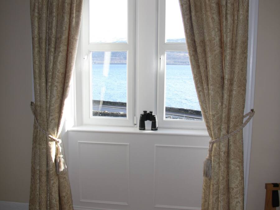 A room with a large window, beige curtains, and a view of the sea.