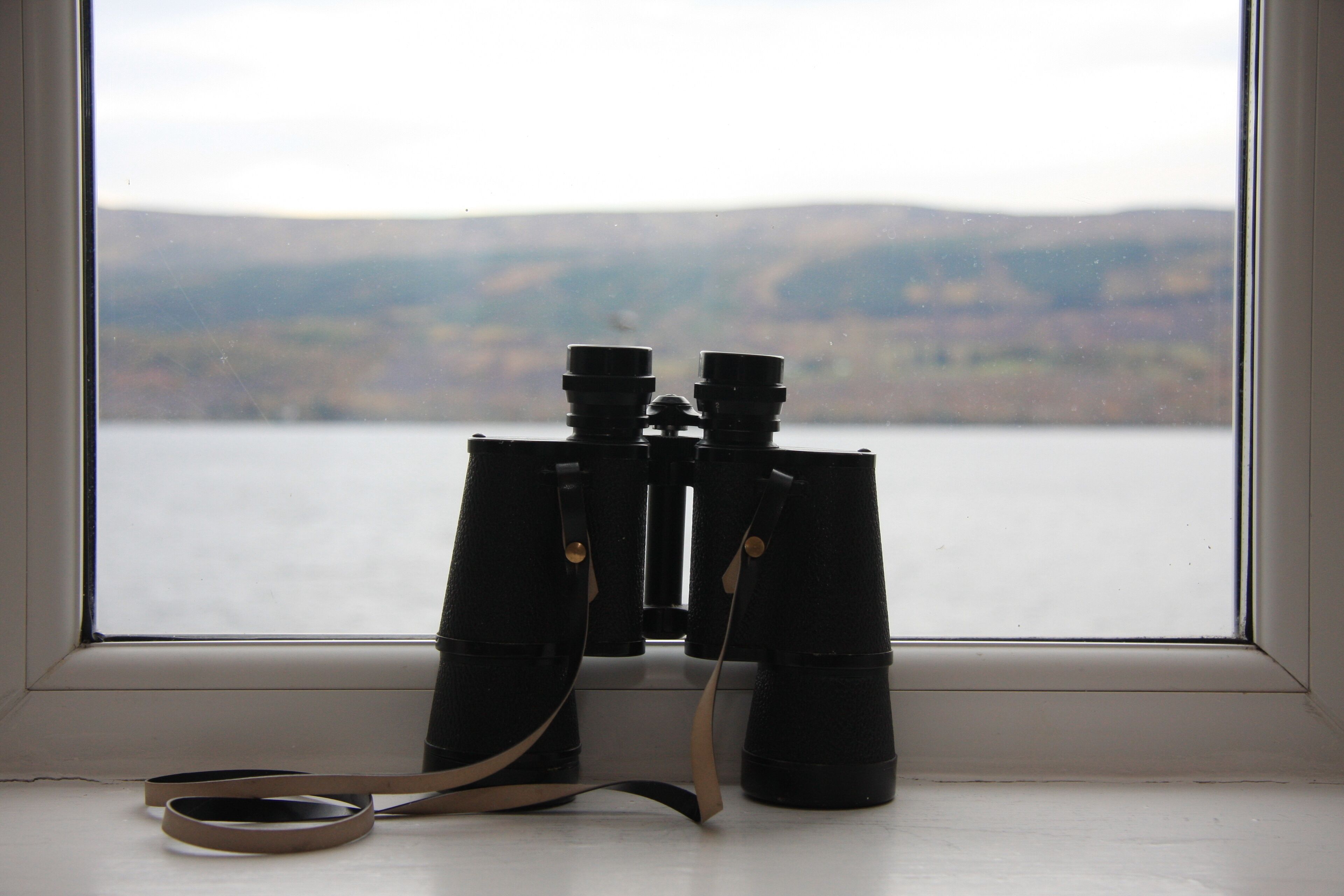 A pair of binoculars placed on a windowsill with a view of a lake and distant hills.