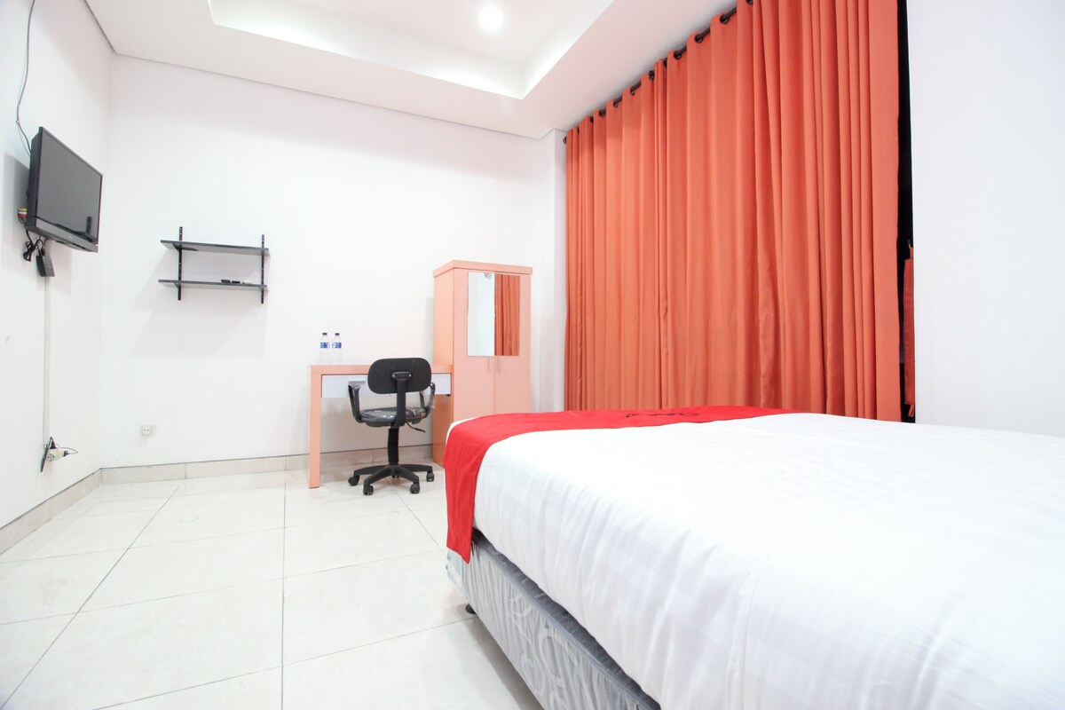 A bedroom with a bed, a desk with a chair, a TV mounted on the wall, and a window with curtains.