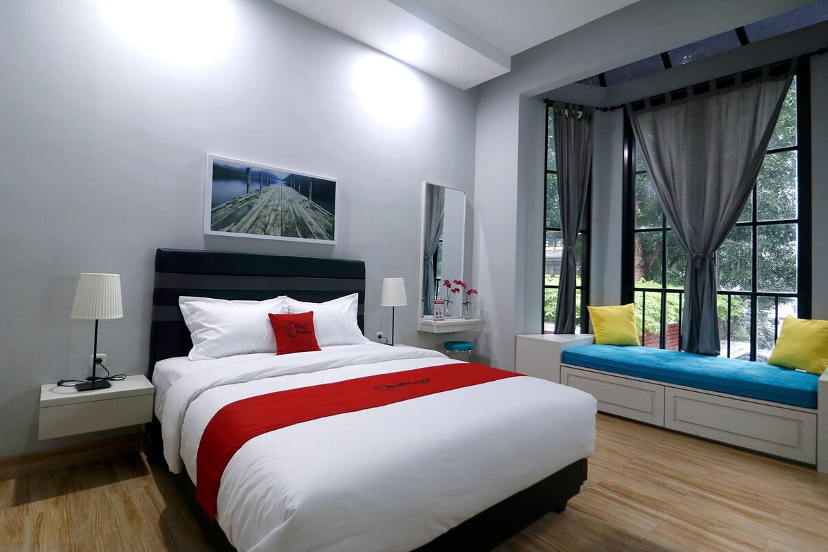 A modern hotel room with a large bed, bedside tables, a window seat, and a view of greenery.
