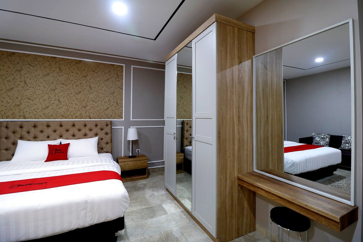 A hotel room with a bed, bedside table, lamp, wardrobe, mirror, and a stool.