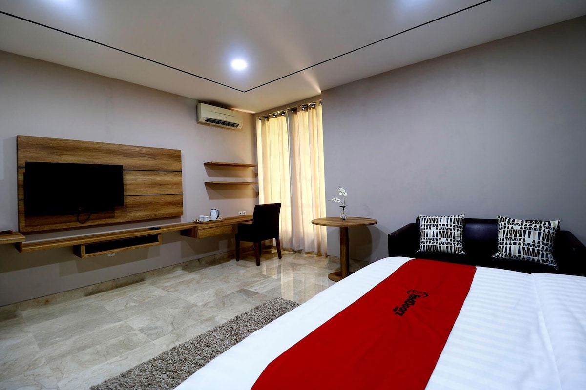 A hotel room with a bed, a TV mounted on the wall, a desk with a chair, a small table with a vase, and a window with curtains.