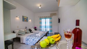 Una habitación de hotel con cama, una mesa de comedor con frutas y una botella, y un ventanal con cortinas.