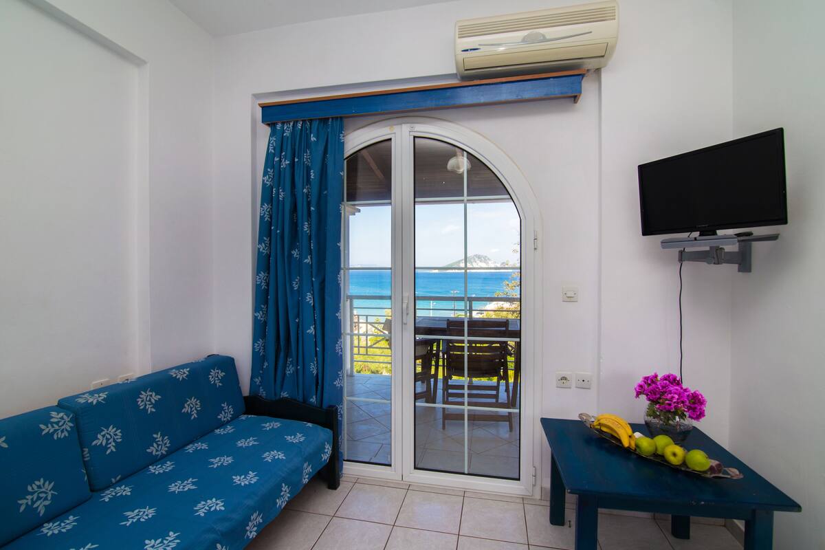 A room with a sofa, a table with fruit and flowers, a TV, and a view of the sea through a glass door.
