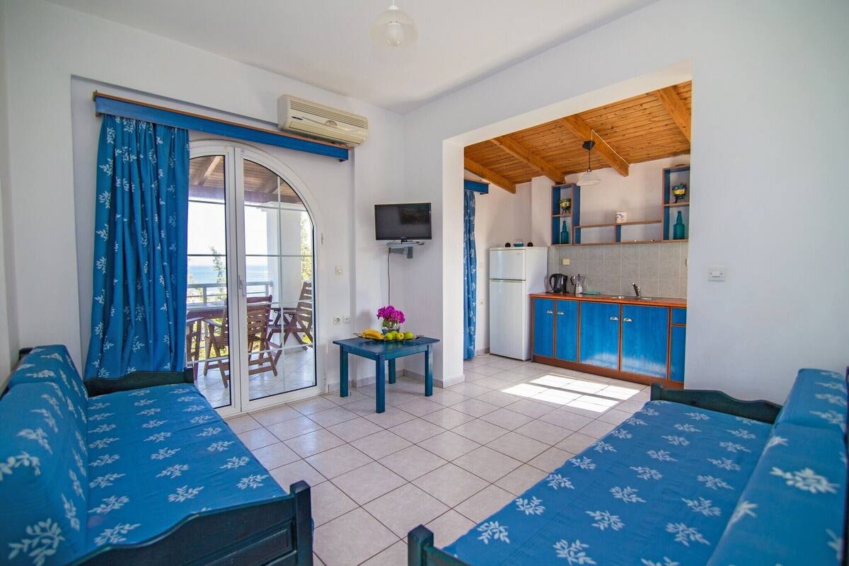 A room with a sofa, a small table with a vase, a television, and a kitchen area with cabinets and a sink.