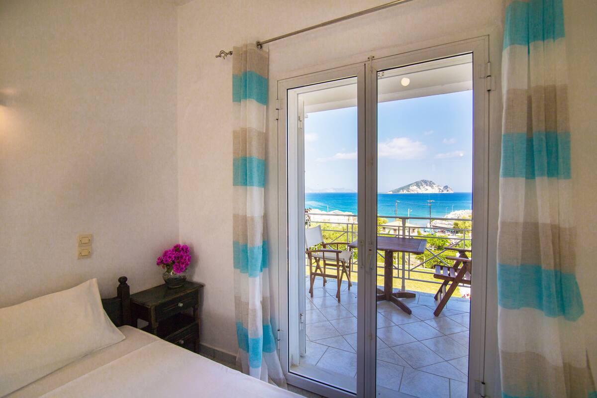 A bedroom with a bed, a nightstand with a vase of flowers, and a view of the sea through a sliding glass door.