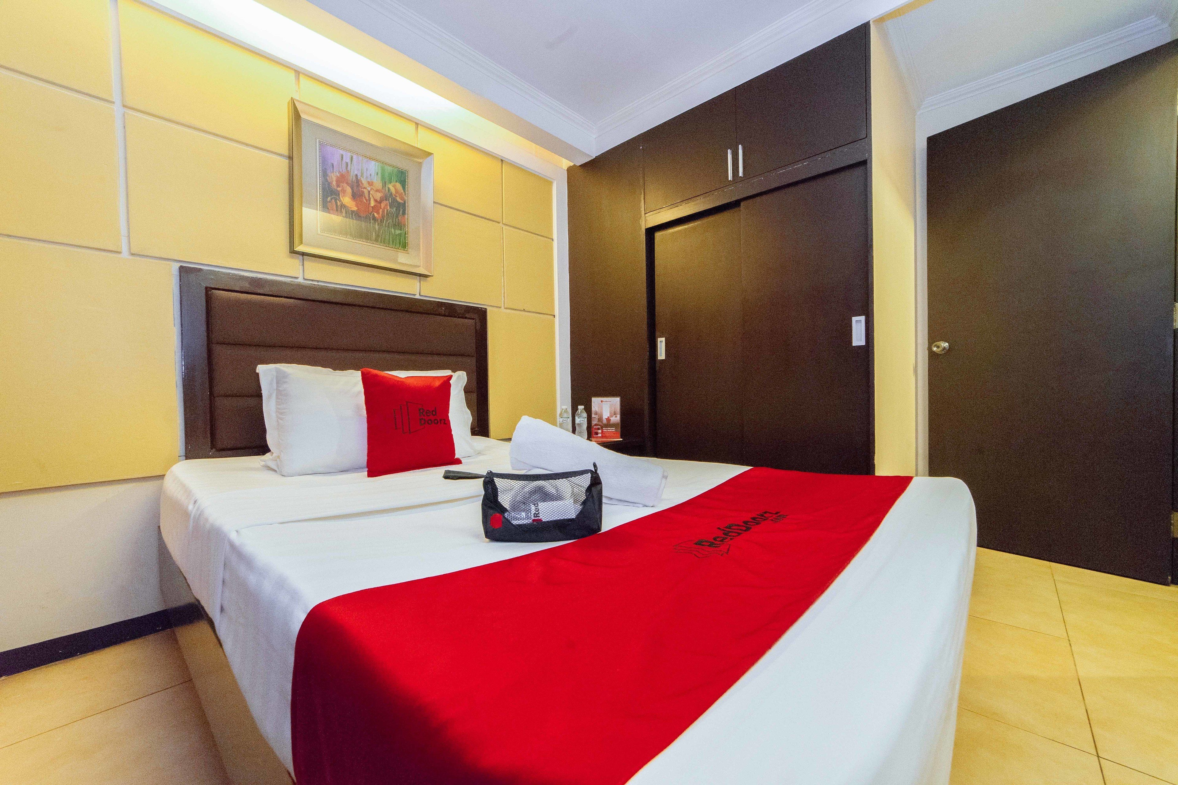 A hotel room with a bed, a red and white bedspread, a red pillow, a white pillow, a headboard, a framed picture on the wall, and a door.