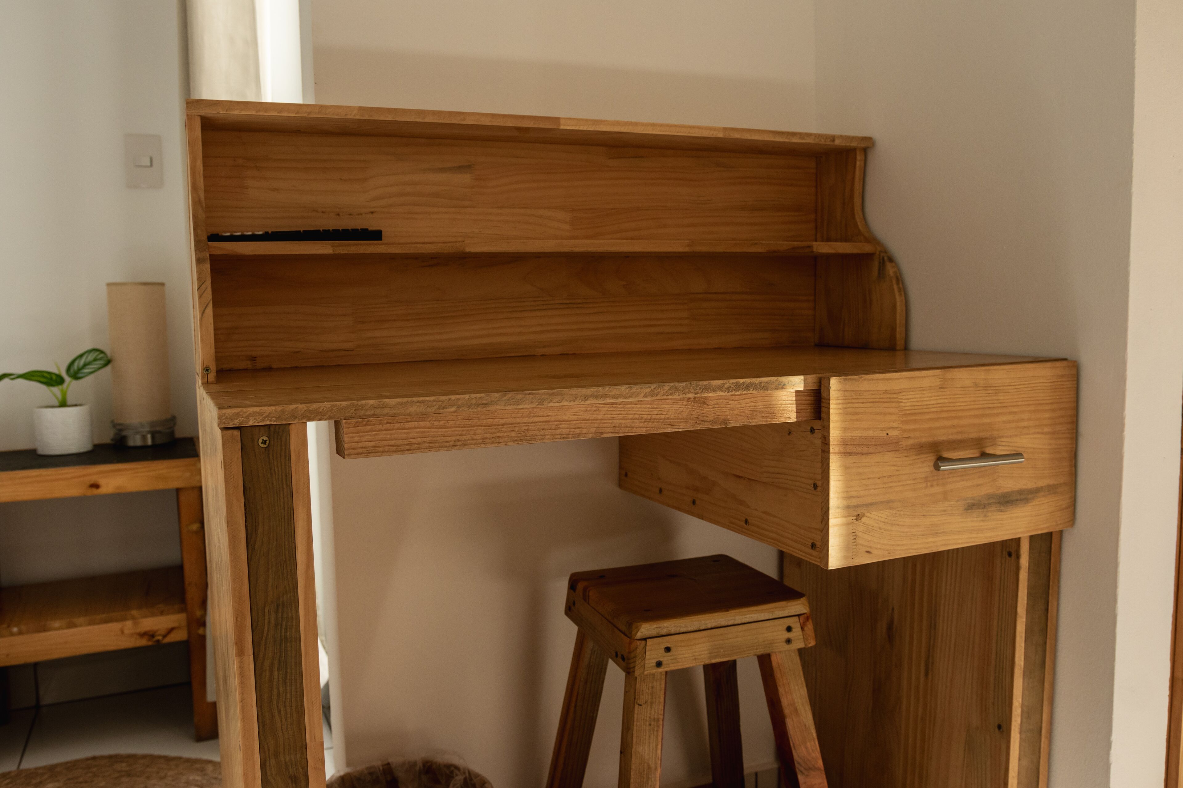 Un escritorio de madera con un cajón y una repisa, un taburete pequeño y una planta en maceta sobre una mesa de madera.