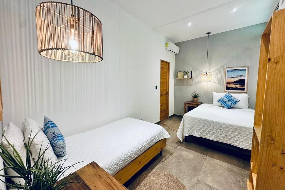 A bedroom with two beds, a wooden headboard, a hanging light fixture, a small bedside table, and a wall-mounted air conditioning unit.