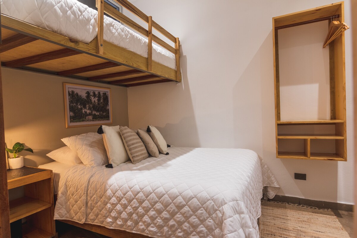 A bedroom with a bunk bed, a bedside table with a plant, a framed picture on the wall, and a wooden shelf unit.