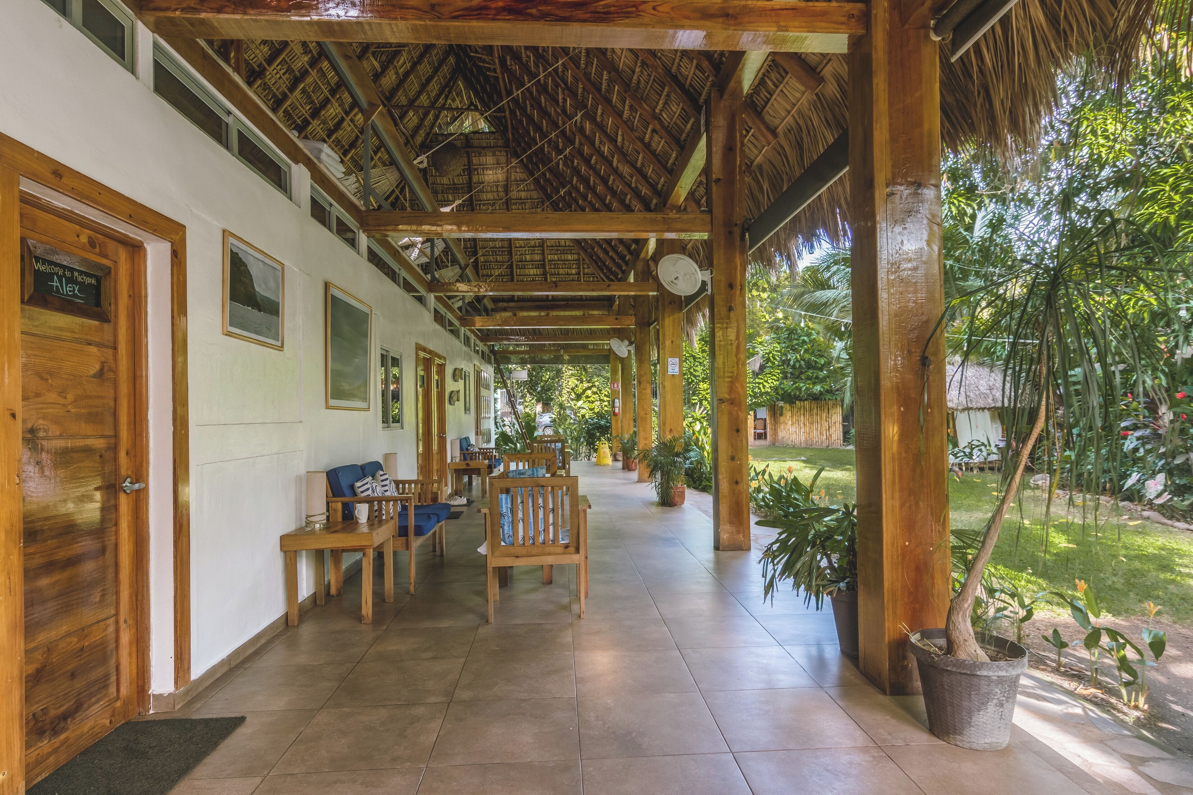 A covered outdoor seating area with wooden benches and tables, surrounded by potted plants and a thatched roof structure.