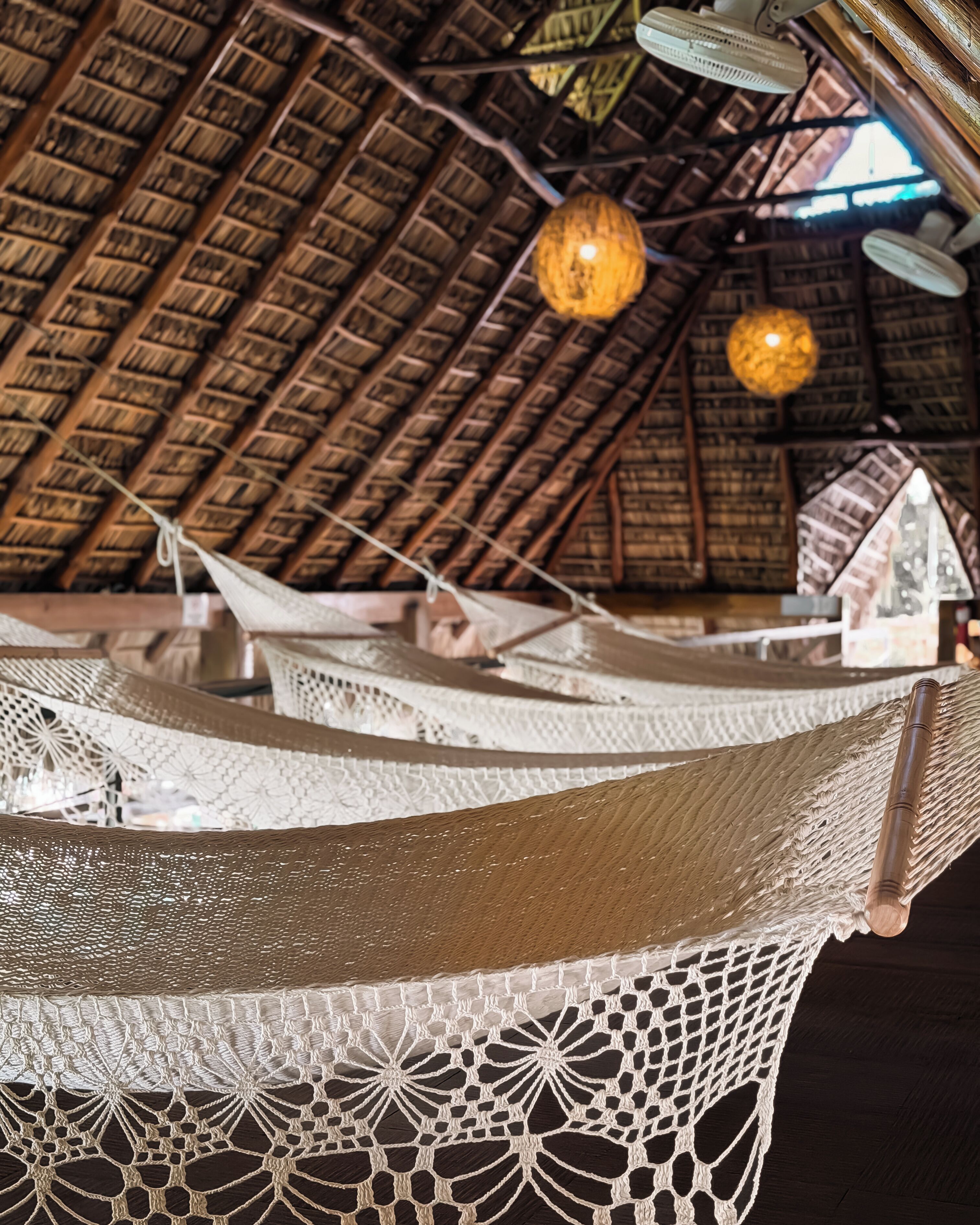 A hammock with a white netted bottom, a wooden ceiling with a thatched roof, and hanging light fixtures.