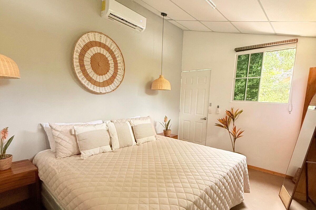A bedroom with a large bed, a ceiling fan, an air conditioning unit, a window with a view, and a door.