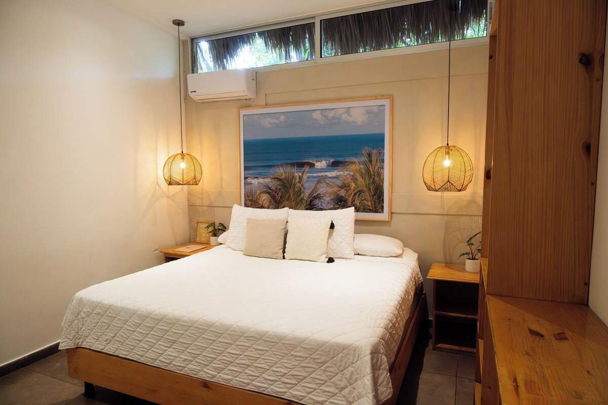 A bedroom with a large bed, a painting of a beach scene, two bedside lamps, and a wooden nightstand.