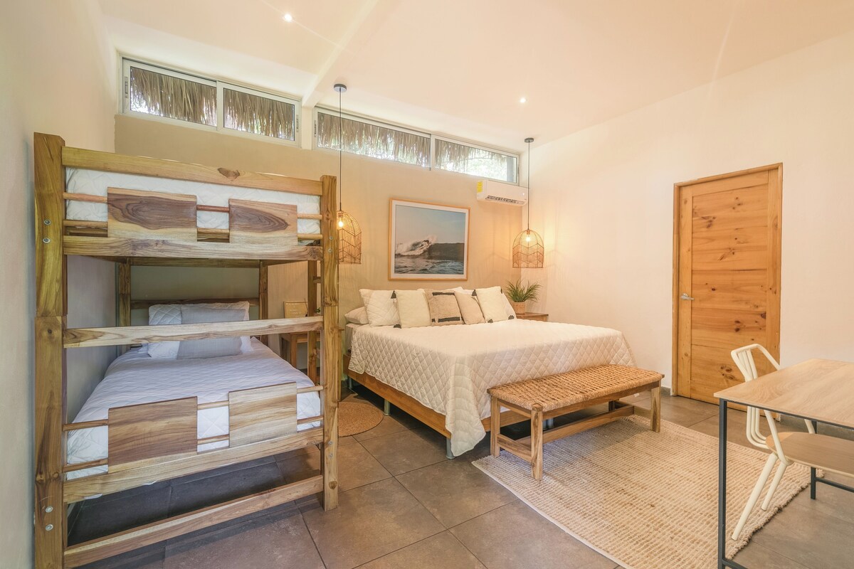 A room with a bunk bed, a single bed, a wooden bench, a table, and a door.