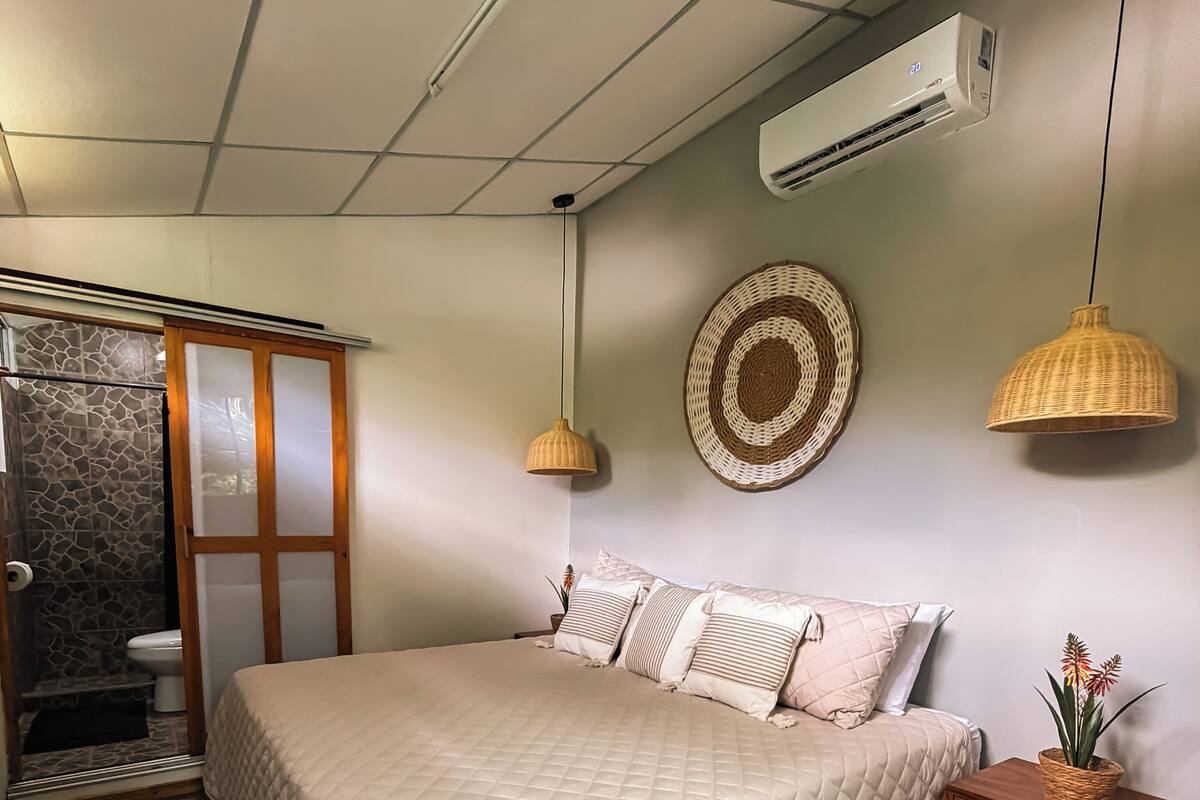 A bedroom with a bed, a wooden nightstand, a wall-mounted air conditioner, a woven basket with flowers, and a woven wall hanging.