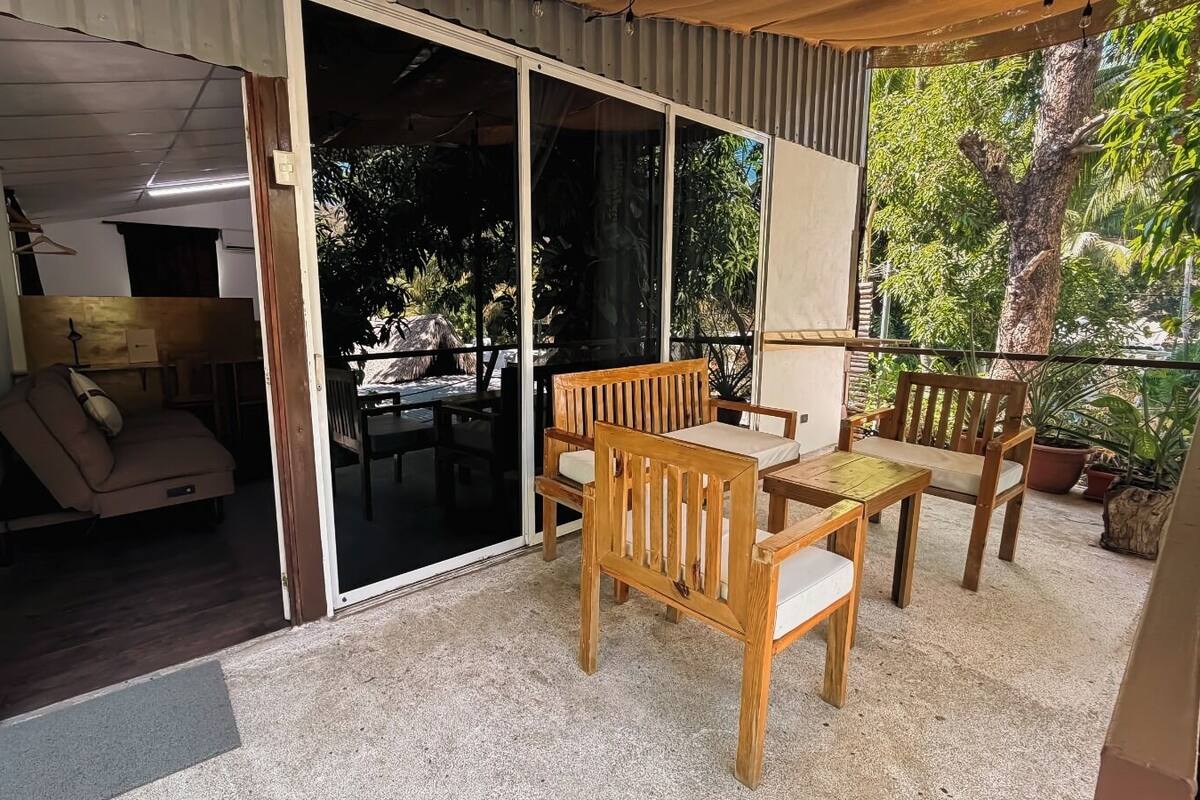 A covered outdoor area with a sliding glass door leading to a room, wooden furniture, and potted plants.