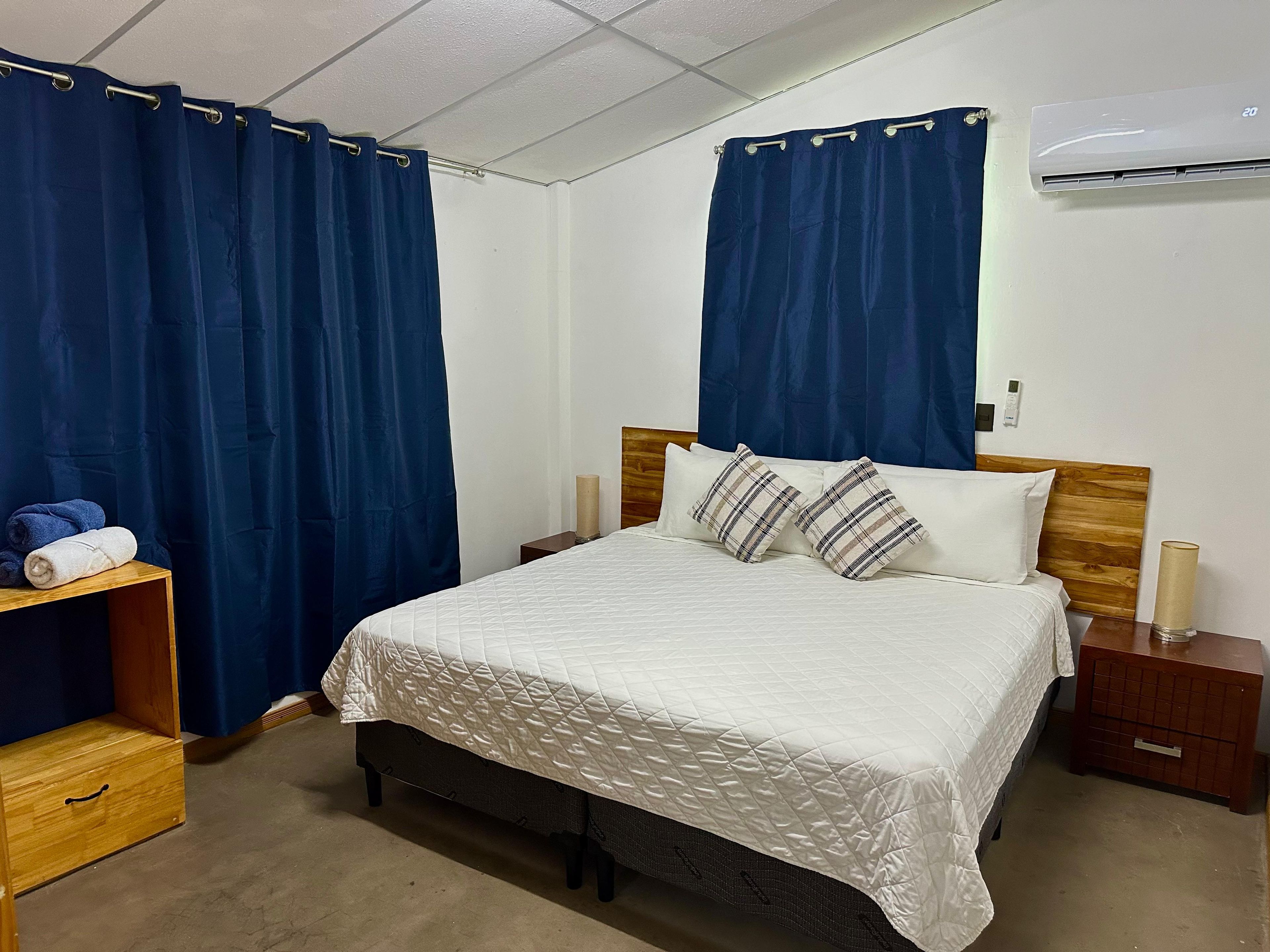 A hotel room with a bed, curtains, a bedside table, a nightstand, and an air conditioning unit.
