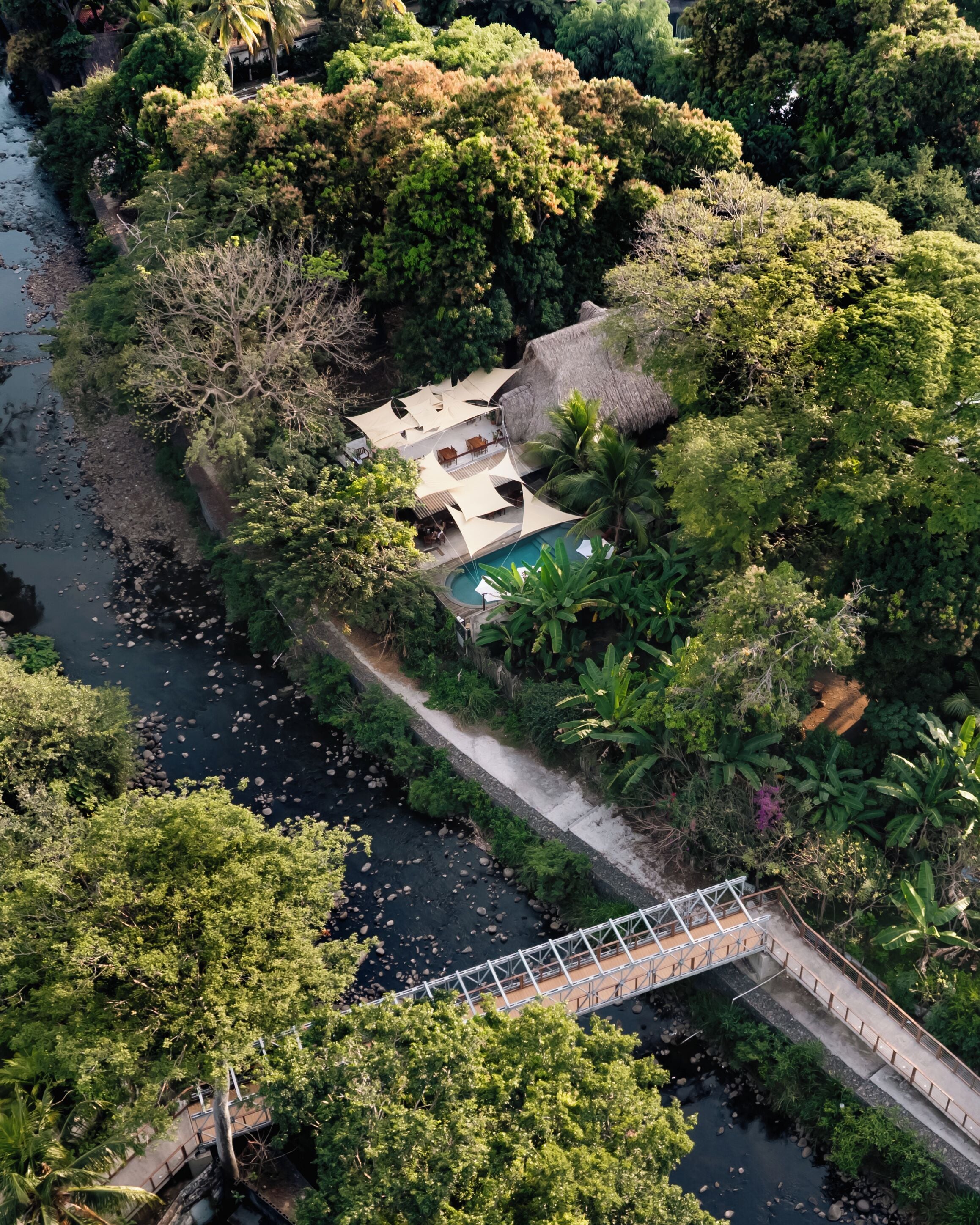 An aerial view of a resort with a swimming pool, a thatched-roof structure, and a bridge over a river.