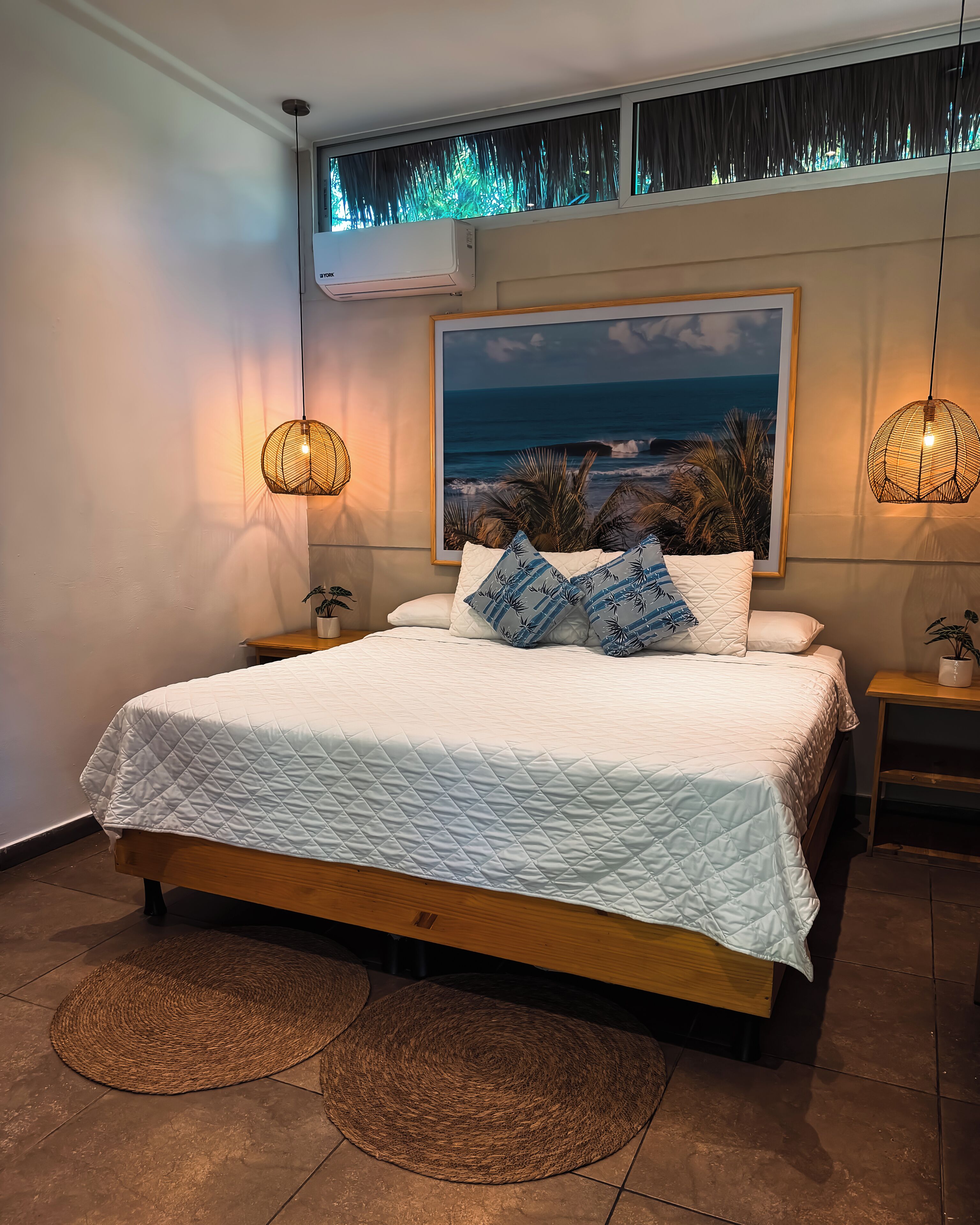 A bedroom with a bed, two bedside tables, a wall-mounted air conditioner, a framed picture, and two hanging lamps.