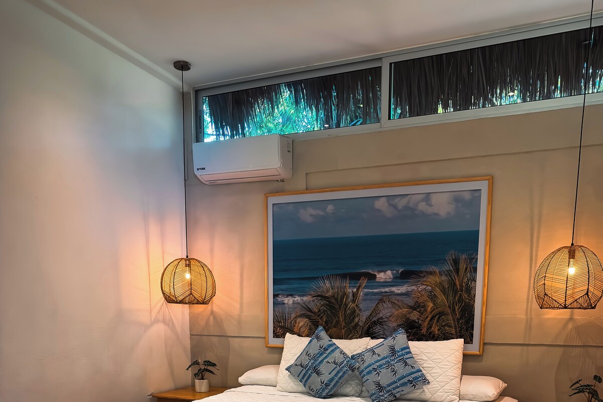 A bedroom with a bed, two bedside tables, a wall-mounted air conditioner, a framed picture, and two hanging lamps.