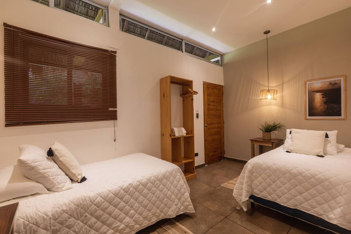 A bedroom with two beds, a wooden wardrobe, a bedside table with a plant, and a painting on the wall.